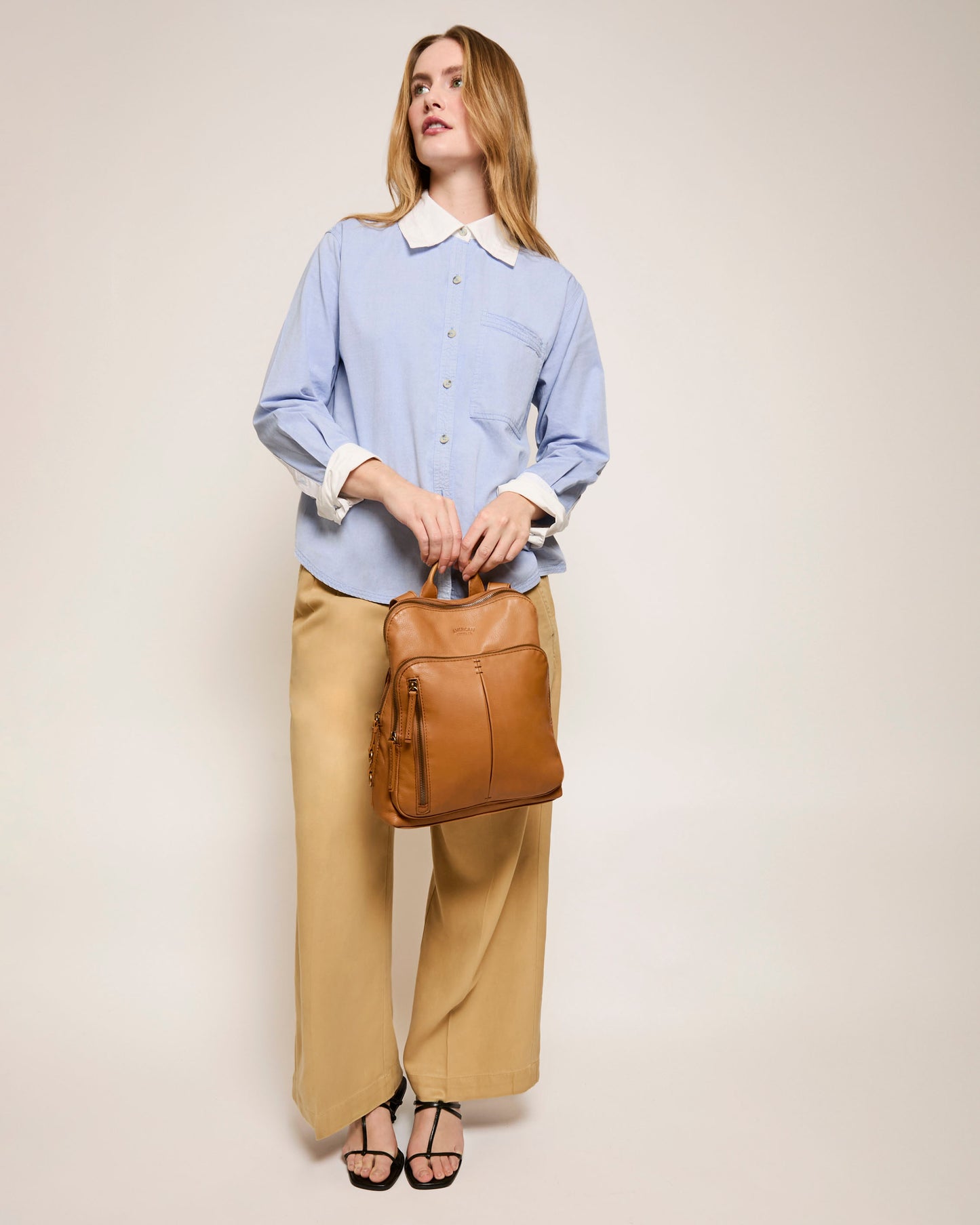 A woman stands against a plain background in a light blue button-up shirt and beige wide-leg pants, holding the Cleveland Backpack from American Leather Co. by its top handles, showcasing its adjustable back straps. She completes her look with black sandals.