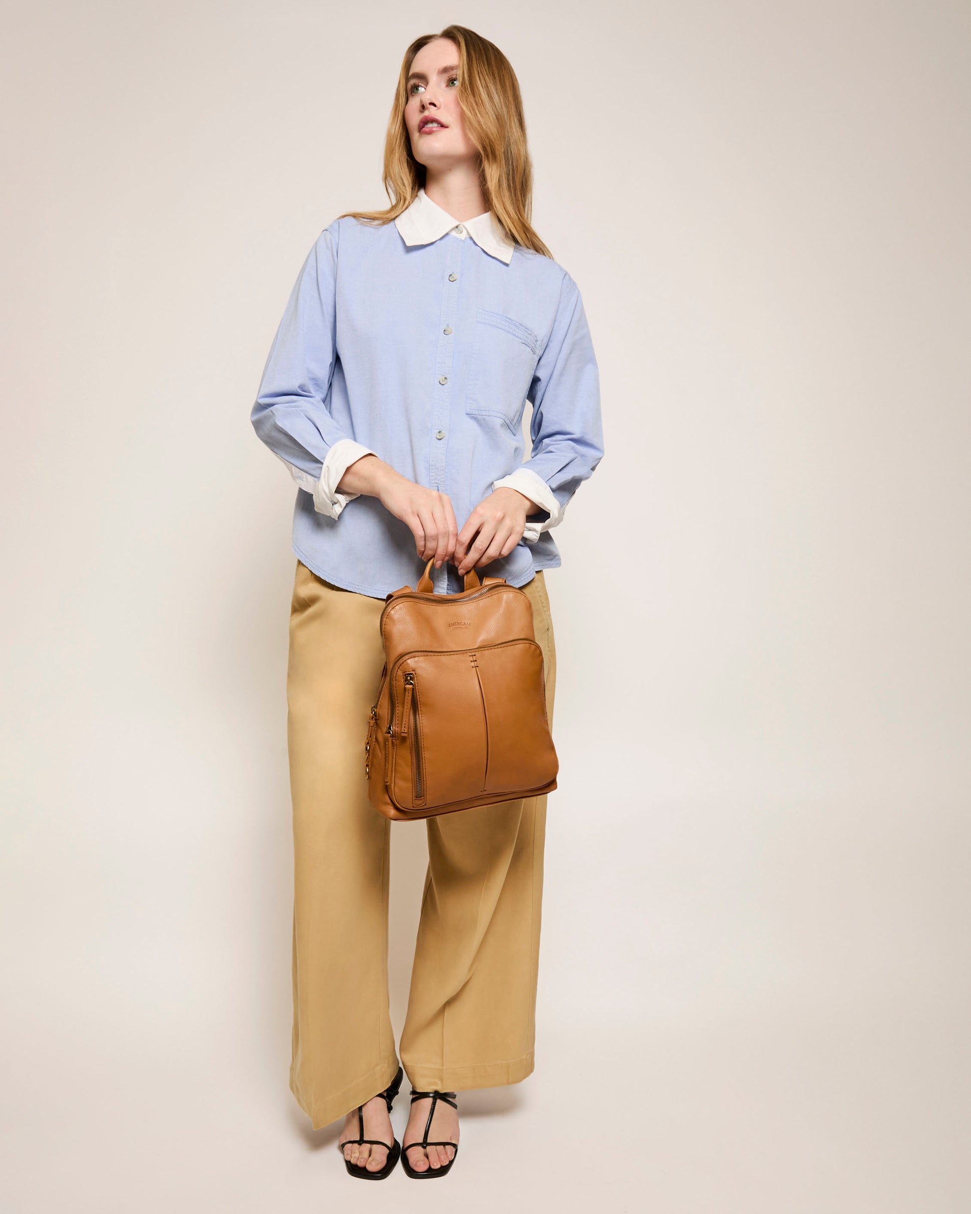 A woman stands against a plain background in a light blue button-up shirt and beige wide-leg pants, holding the Cleveland Backpack from American Leather Co. by its top handles, showcasing its adjustable back straps. She completes her look with black sandals.