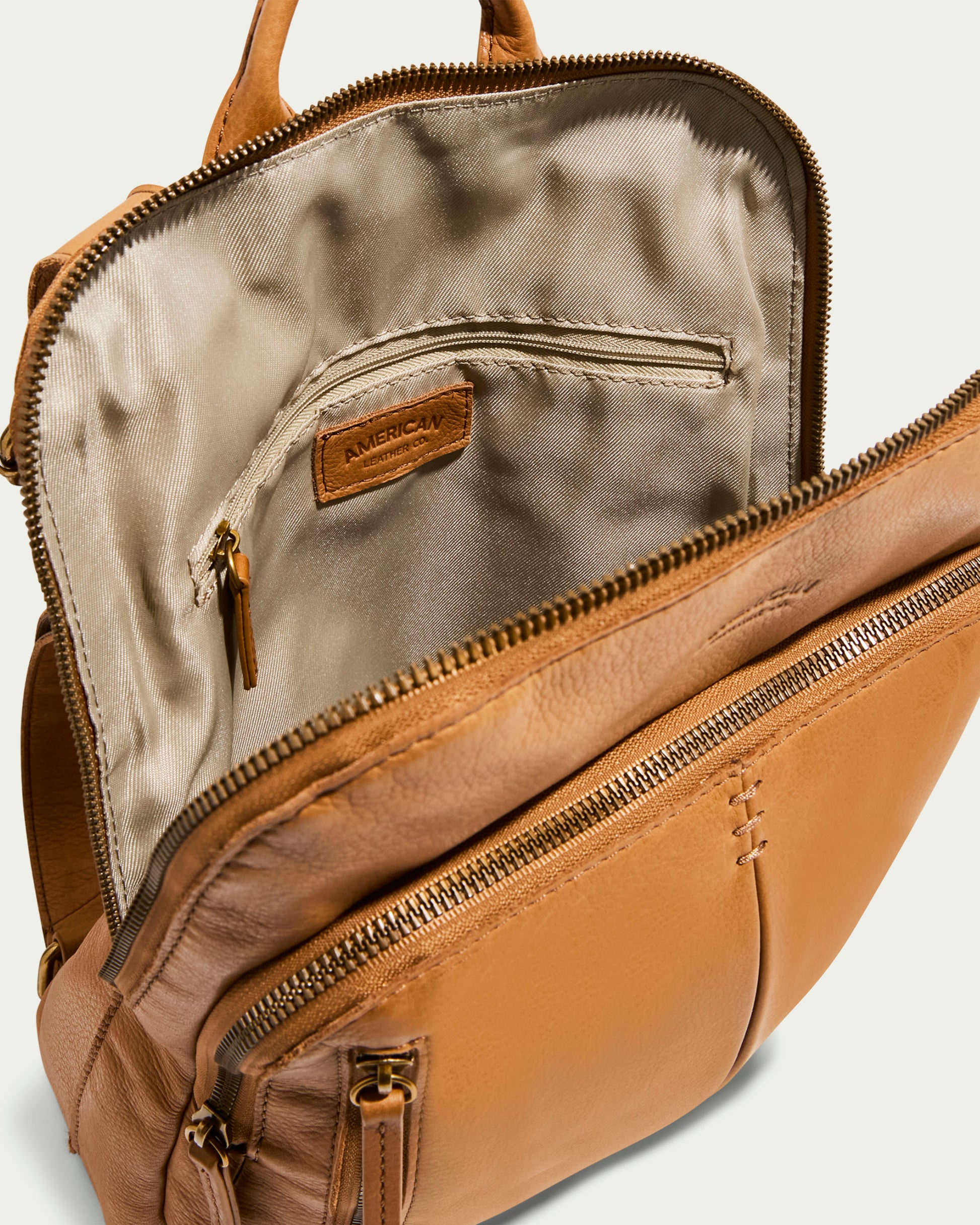 The Cleveland Backpack by American Leather Co. is an open tan women's leather backpack made from genuine American leather, with a front pocket, beige interior lining, zipper closure, inner pocket with a stitched label, and adjustable back straps for comfort.