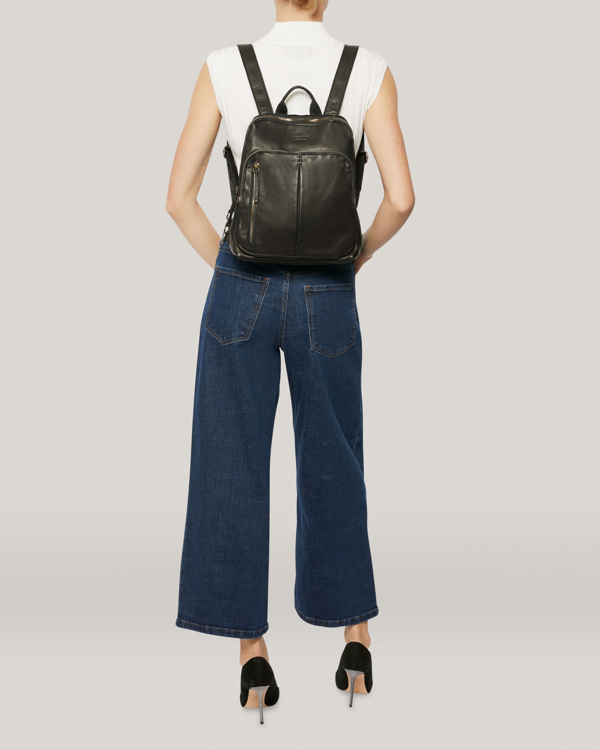 A person is standing with their back to the camera, wearing the black Cleveland Backpack by American Leather Co., which features buttery soft leather and adjustable back straps. They are also dressed in a sleeveless white top, blue wide-leg jeans, and black high-heeled shoes. The background is plain and light-colored.