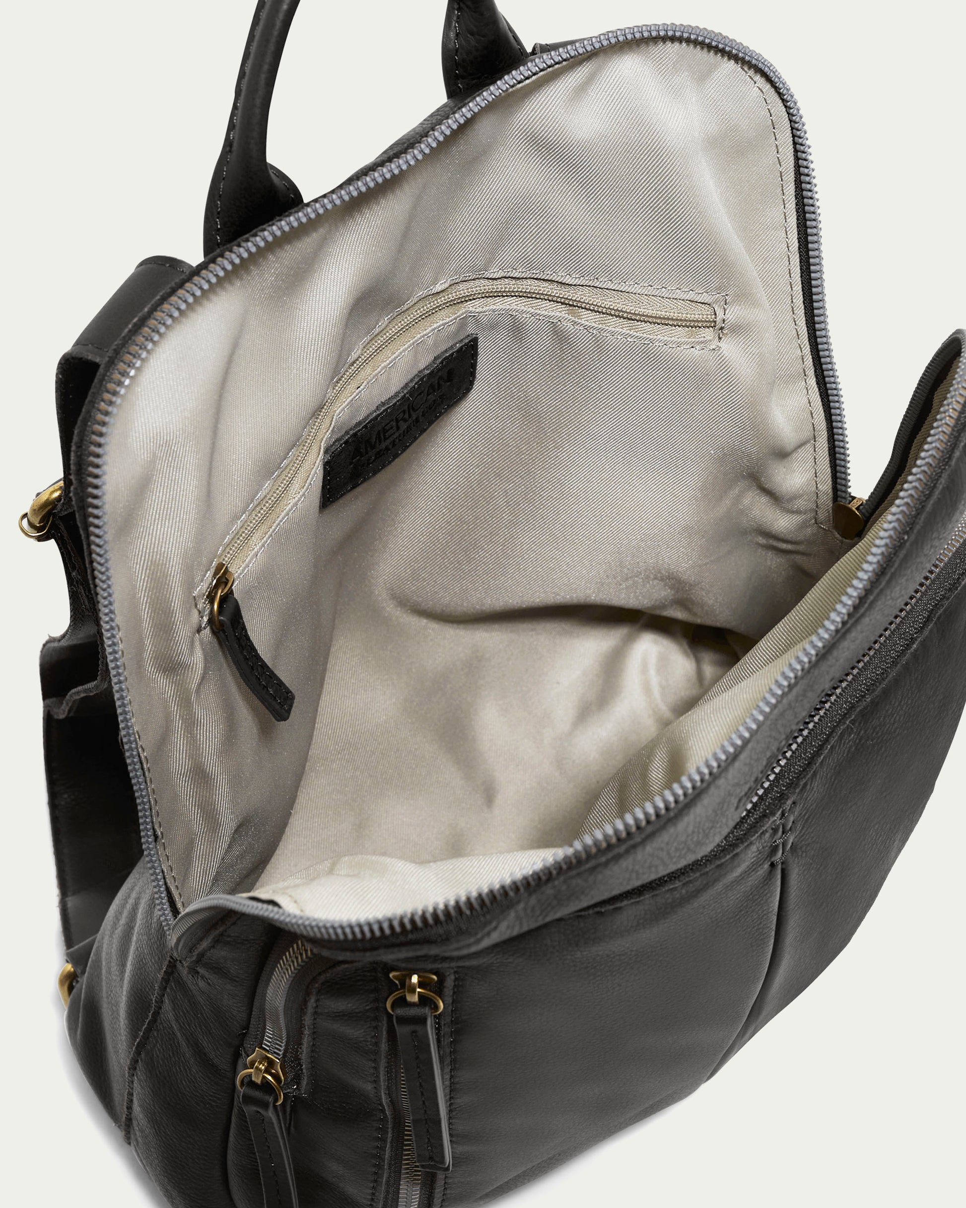 The Cleveland Backpack by American Leather Co. features an open buttery soft leather design that reveals a spacious beige interior with a zippered pocket. The top of the bag is kept open with a U-shaped zipper, showcasing additional pockets and gold-colored zip closures on the outside. Adjustable back straps ensure comfort and versatility.
