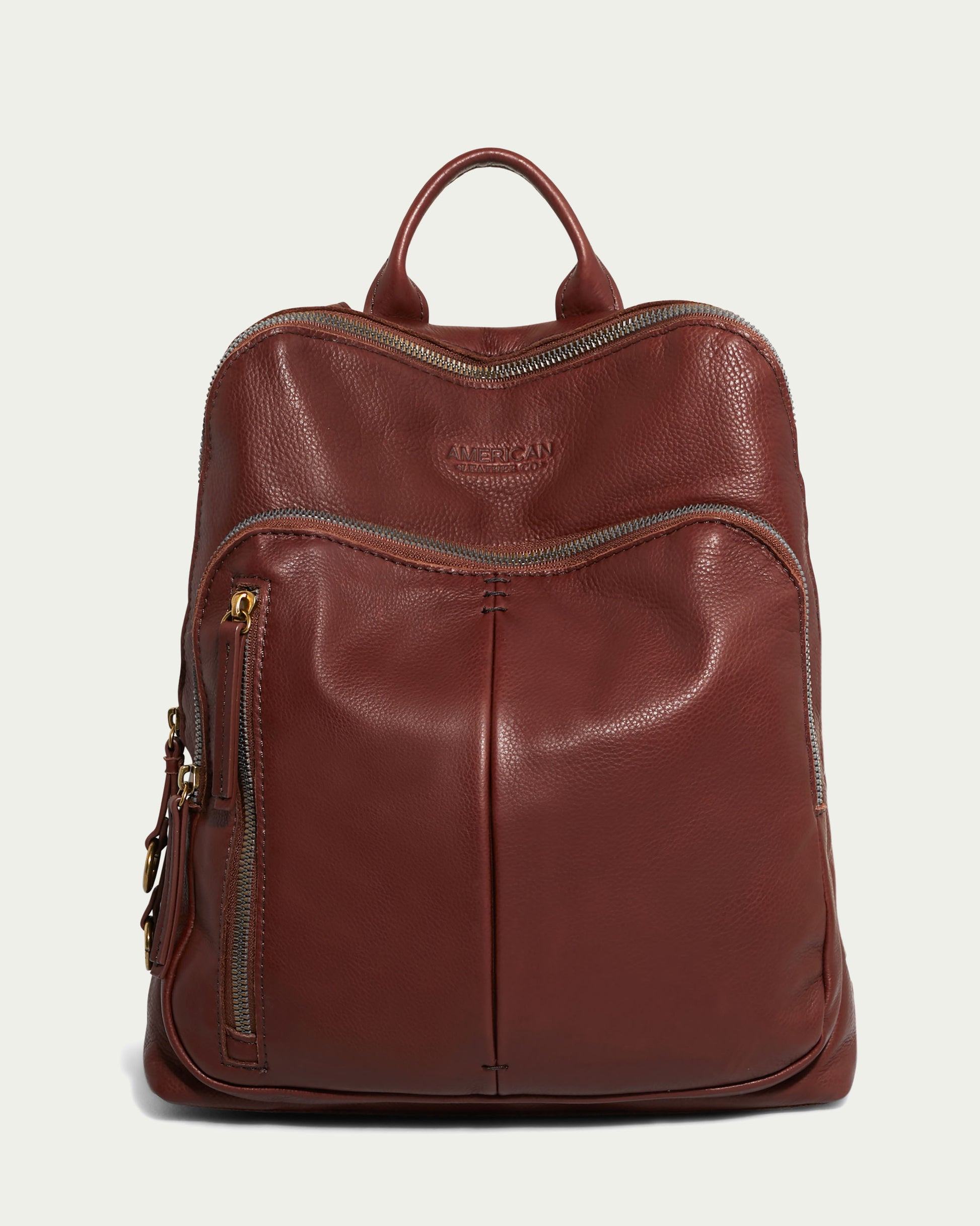 Introducing the Cleveland Backpack by American Leather Co., a brown leather backpack purse crafted from genuine American leather. This stylish accessory features a front zippered pocket, two vertical seams, adjustable back straps, and a top handle. The main compartment secures with a zip closure, while the brand logo embossed on the upper front combines elegantly with gold-tone hardware for added sophistication.