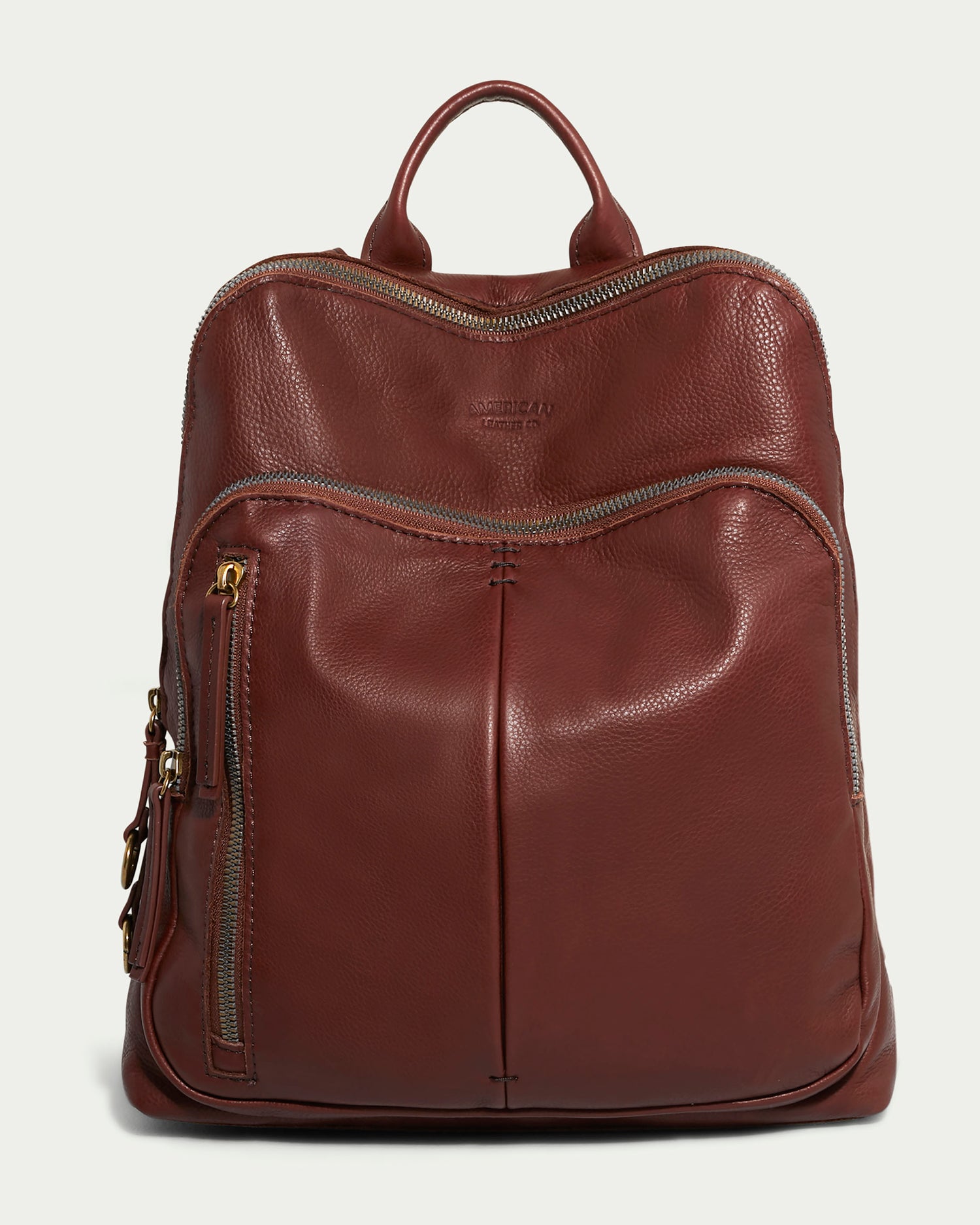 The Cleveland Backpack by American Leather Co. is a brown leather purse with antiqued hardware and silver zippers, featuring a front diagonal zipper pocket and a side vertical zipper pocket. The genuine leather highlights quality, complemented by a functional top handle.