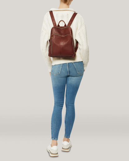 A person is standing with their back to the camera, wearing a white knitted sweater, blue jeans, and white sneakers. They are carrying a Cleveland Backpack by American Leather Co., made of genuine brown American leather with adjustable back straps. The background is plain and light-colored.