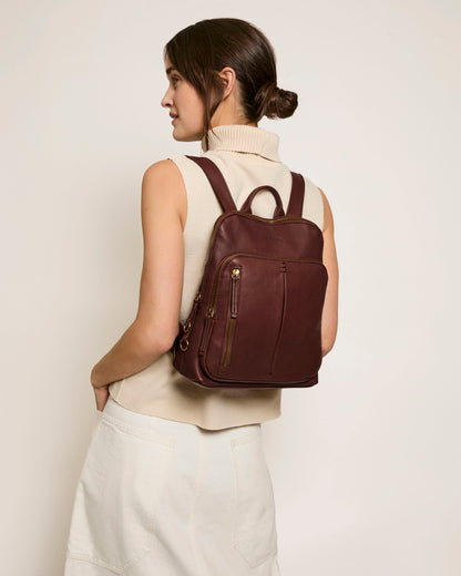 A person with brown hair in a low bun is wearing a sleeveless beige turtleneck and a white skirt. They have the Cleveland Backpack from American Leather Co., crafted from genuine leather, slung over their shoulder, as they look to the side against a plain background.