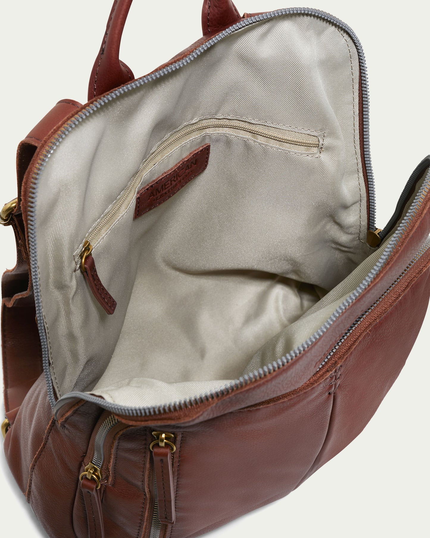 The Cleveland Backpack by American Leather Co. unveils its beige interior lining and inside zippered pocket when opened. This brown leather backpack purse also boasts several external zippered compartments, gold-tone hardware, and adjustable back straps, all intricately crafted from genuine American leather.