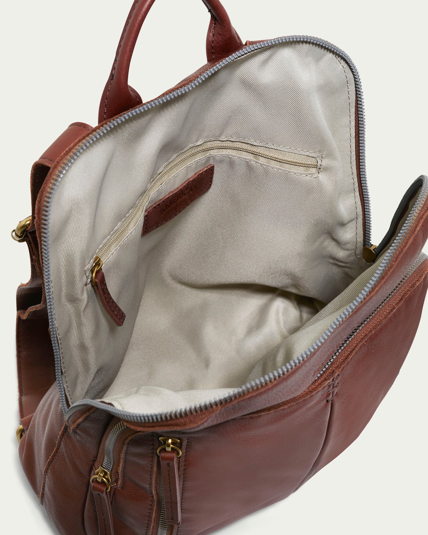 The Cleveland Backpack by American Leather Co. is a brown leather purse with an open top zipper, revealing a spacious beige interior made of genuine American leather. It features an interior side zipper pocket and exterior pockets adorned with antiqued gold-tone hardware.