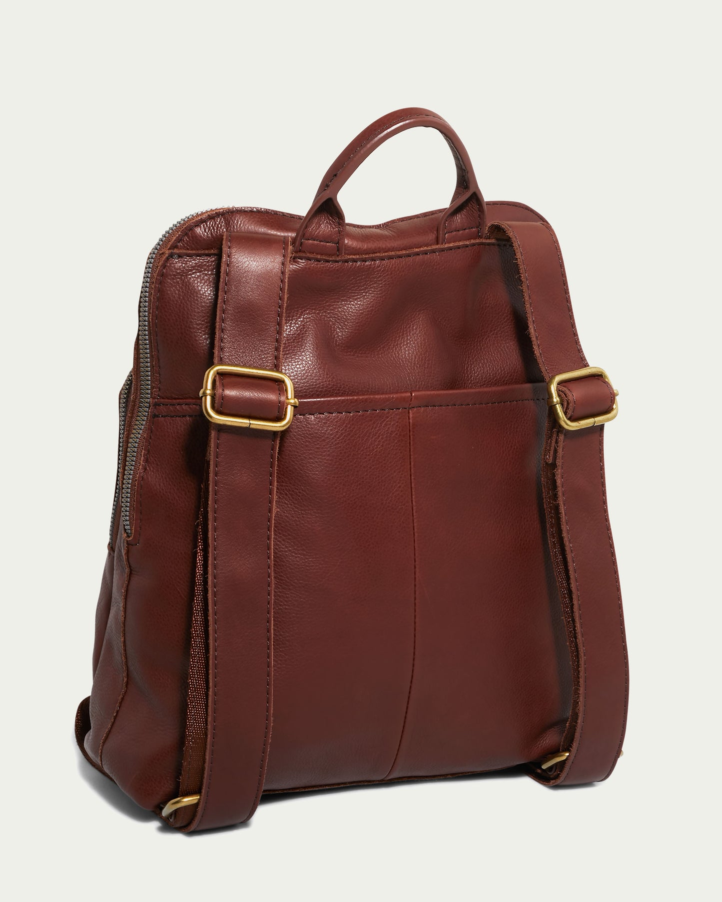 The Cleveland Backpack by American Leather Co. is a genuine leather backpack purse featuring gold-tone buckles on its adjustable shoulder straps. It comes with a top handle, visible stitching, and zippers on each side for easy access to the main compartment. The background is a solid light beige color.