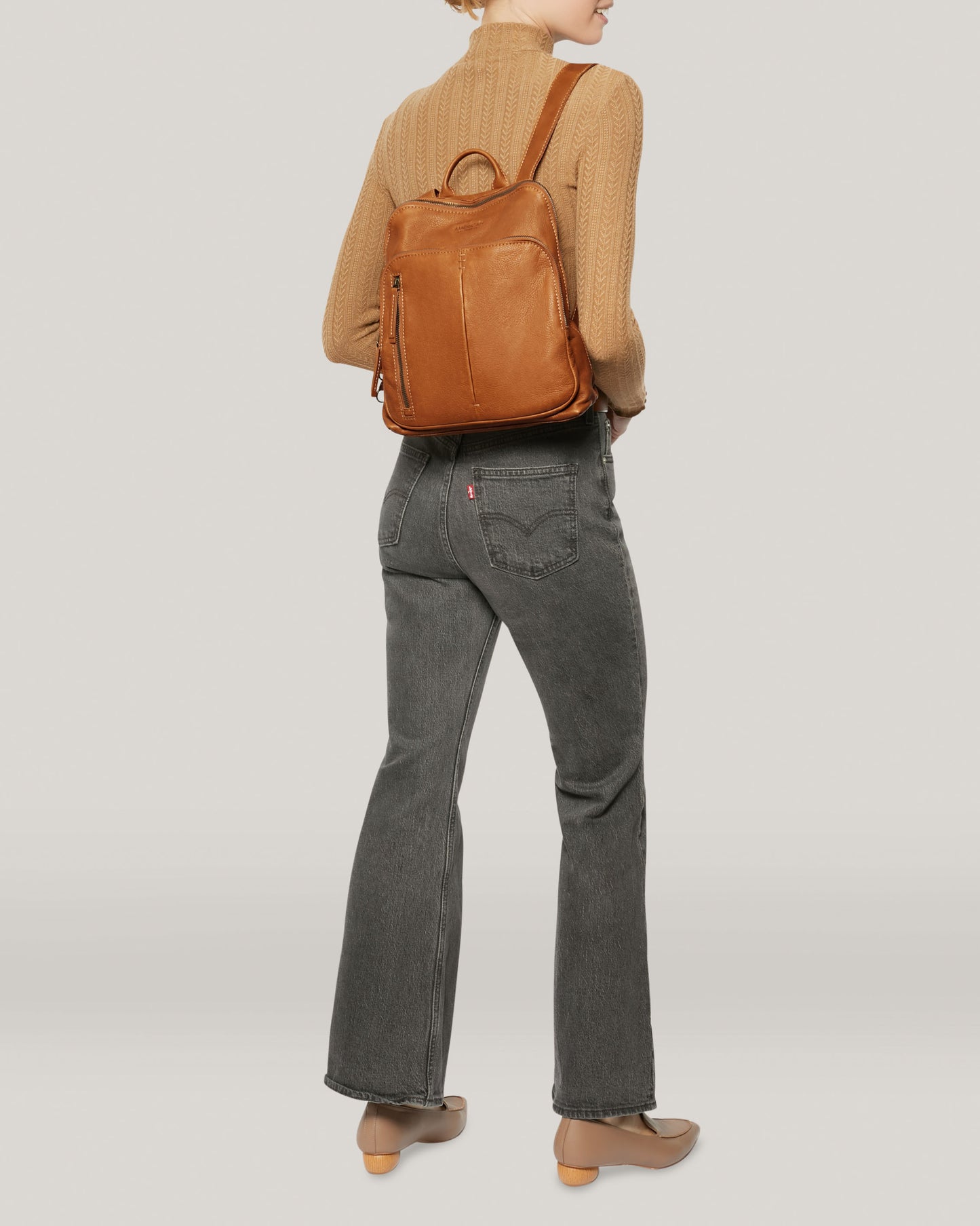 A person wearing a brown sweater, gray jeans, and beige boots is standing with their back to the viewer, highlighting the Cleveland Backpack by American Leather Co., made from buttery soft leather. The background is a simple light gray.