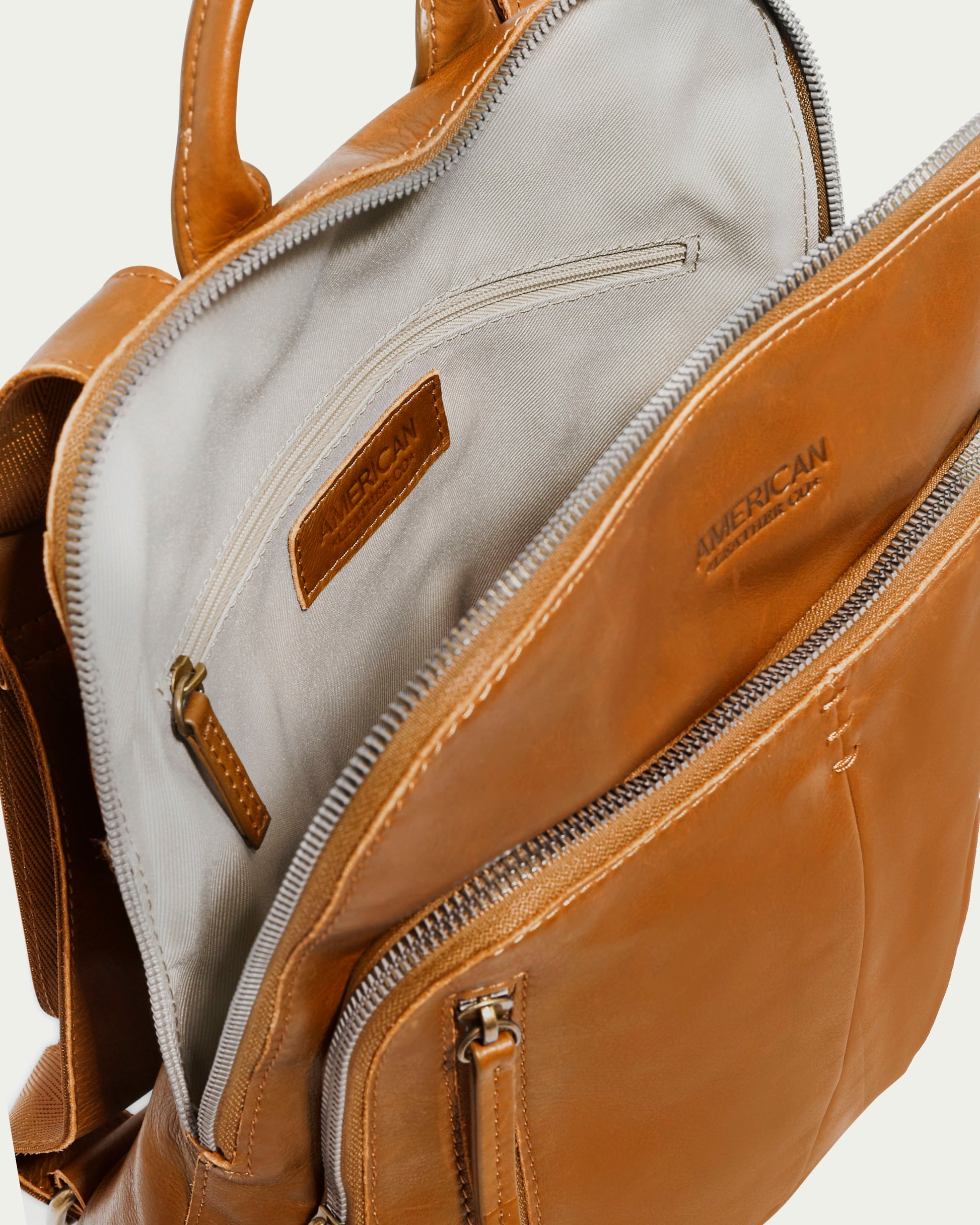 The Cleveland Backpack from American Leather Co. is a tan leather purse featuring multiple compartments and zippers. When open, it reveals a spacious interior lined with light fabric and includes a small zippered pocket. Made from buttery soft leather, the brand name "American Leather Co." is elegantly engraved on the inner label.