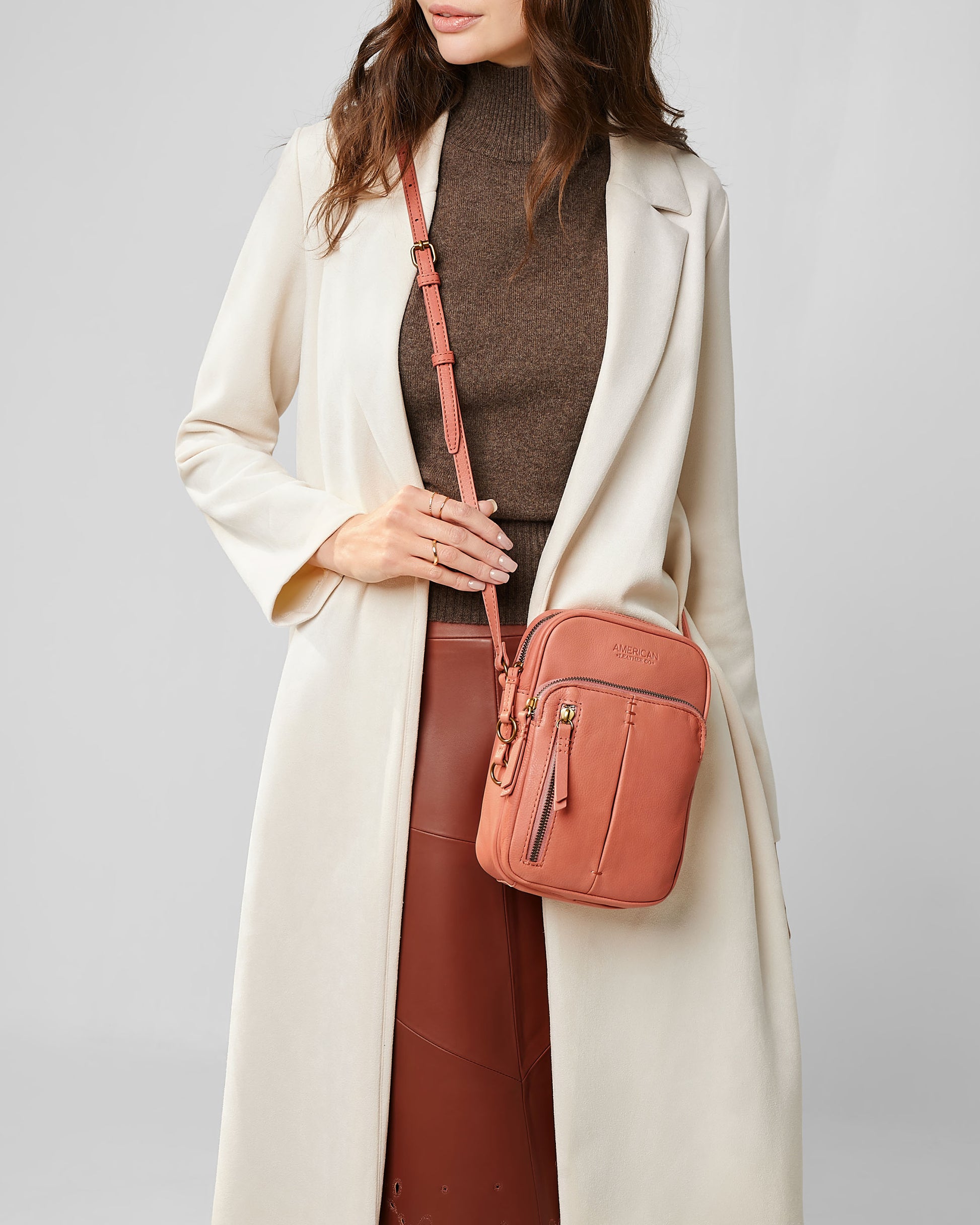 A person wearing a beige coat and brown turtleneck holds the stylish Cleveland Large NS Crossbody bag from American Leather Co., crafted from genuine American leather. The outfit, paired with a brown skirt, is accentuated by the bag's pink color and gold buckle, exuding an elegant style.