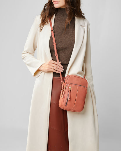 A person wearing a beige coat and brown turtleneck holds the stylish Cleveland Large NS Crossbody bag from American Leather Co., crafted from genuine American leather. The outfit, paired with a brown skirt, is accentuated by the bag's pink color and gold buckle, exuding an elegant style.