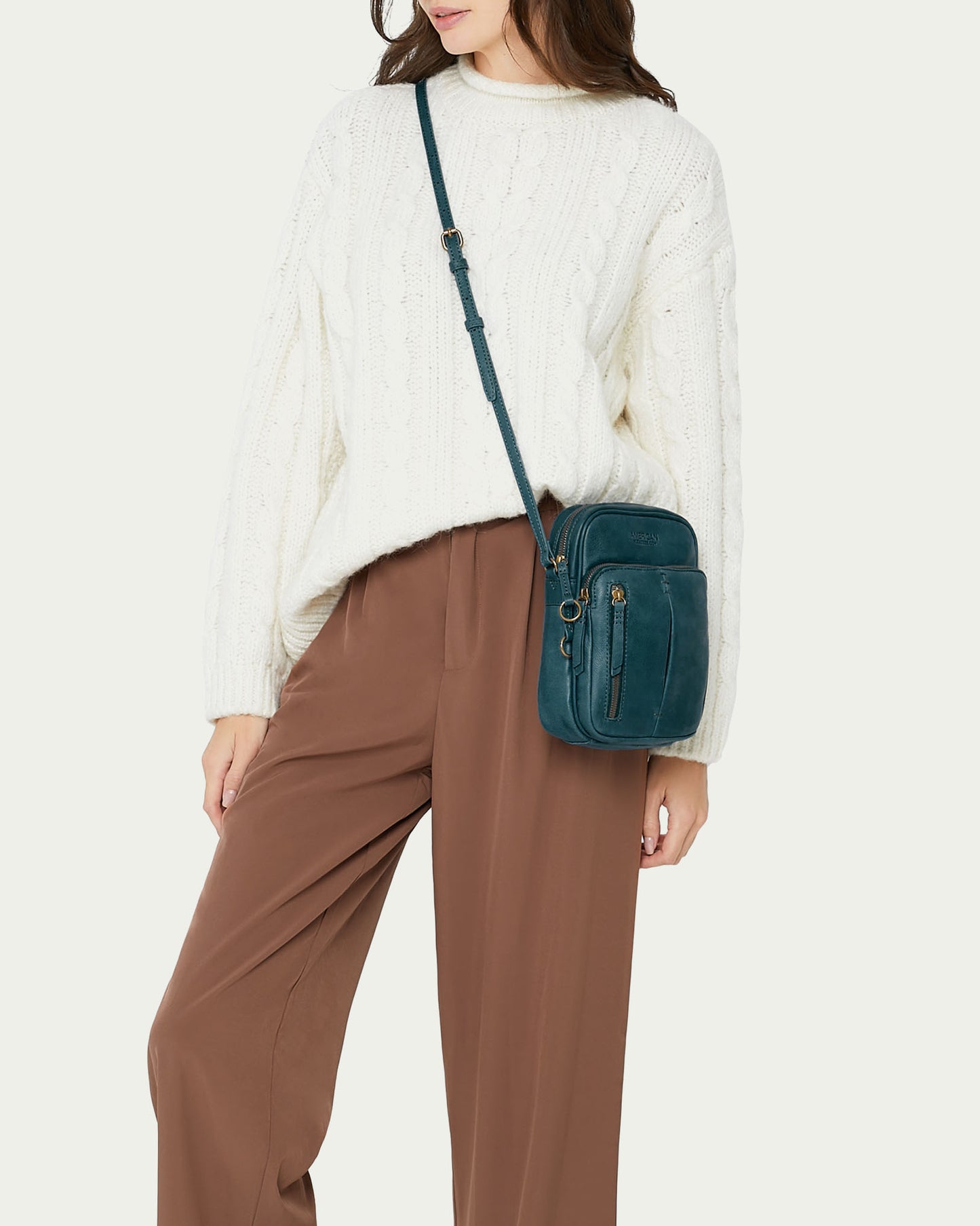 A woman wears a white cable-knit sweater, brown wide-leg pants, and a teal Cleveland Large NS Crossbody bag from American Leather Co.