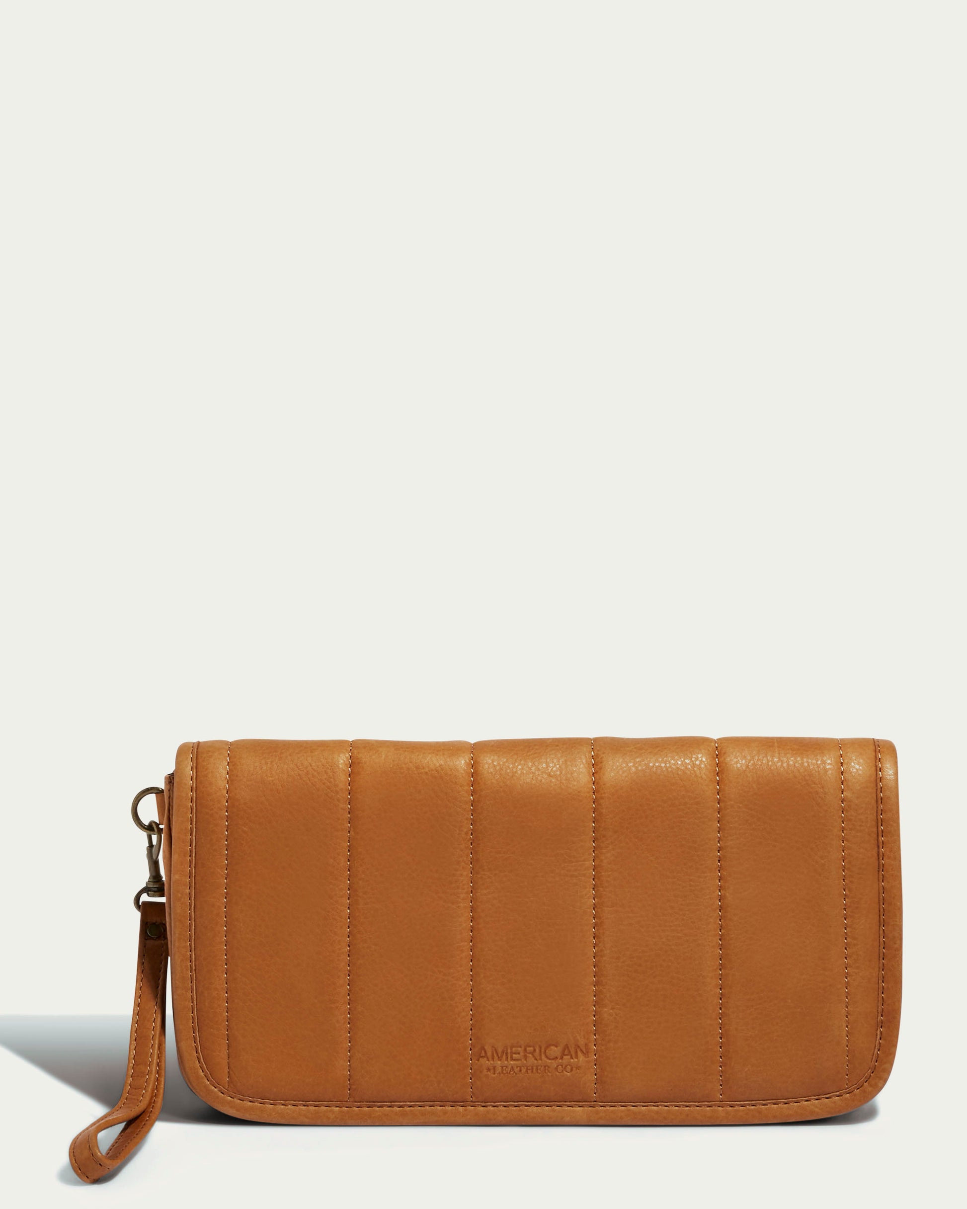 Introducing the Dayton Oversized Clutch by American Leather Co. This tan leather accessory is crafted from glove-tanned leather and showcases vertical panel quilting. It features a detachable wrist strap for versatility, and the brand's name is embossed on the lower front. The background is a light cream color.