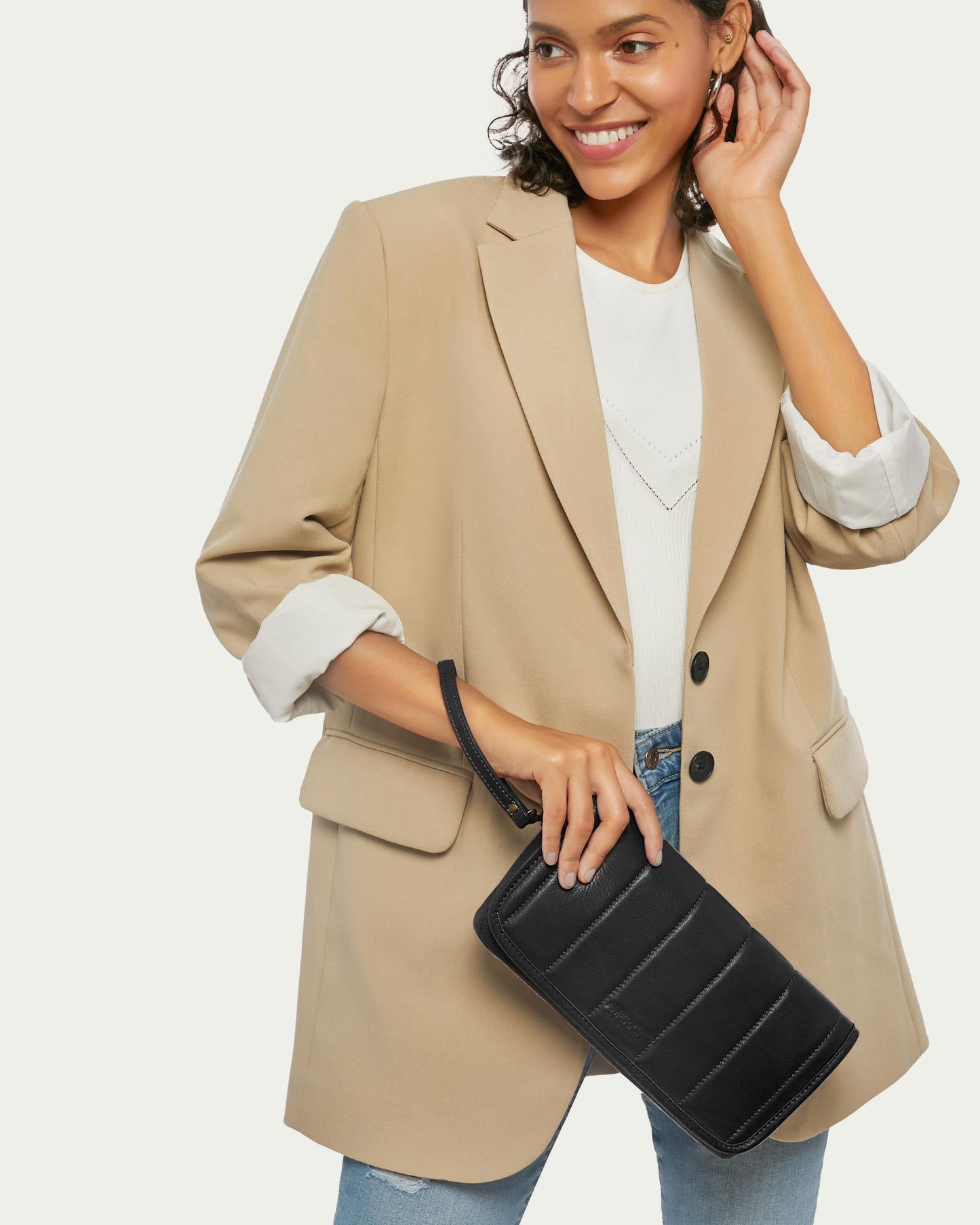 A woman with curly hair smiles while wearing a beige blazer, white top, and jeans. She holds the Dayton Oversized Clutch by American Leather Co., expertly crafted from glove-tanned leather.