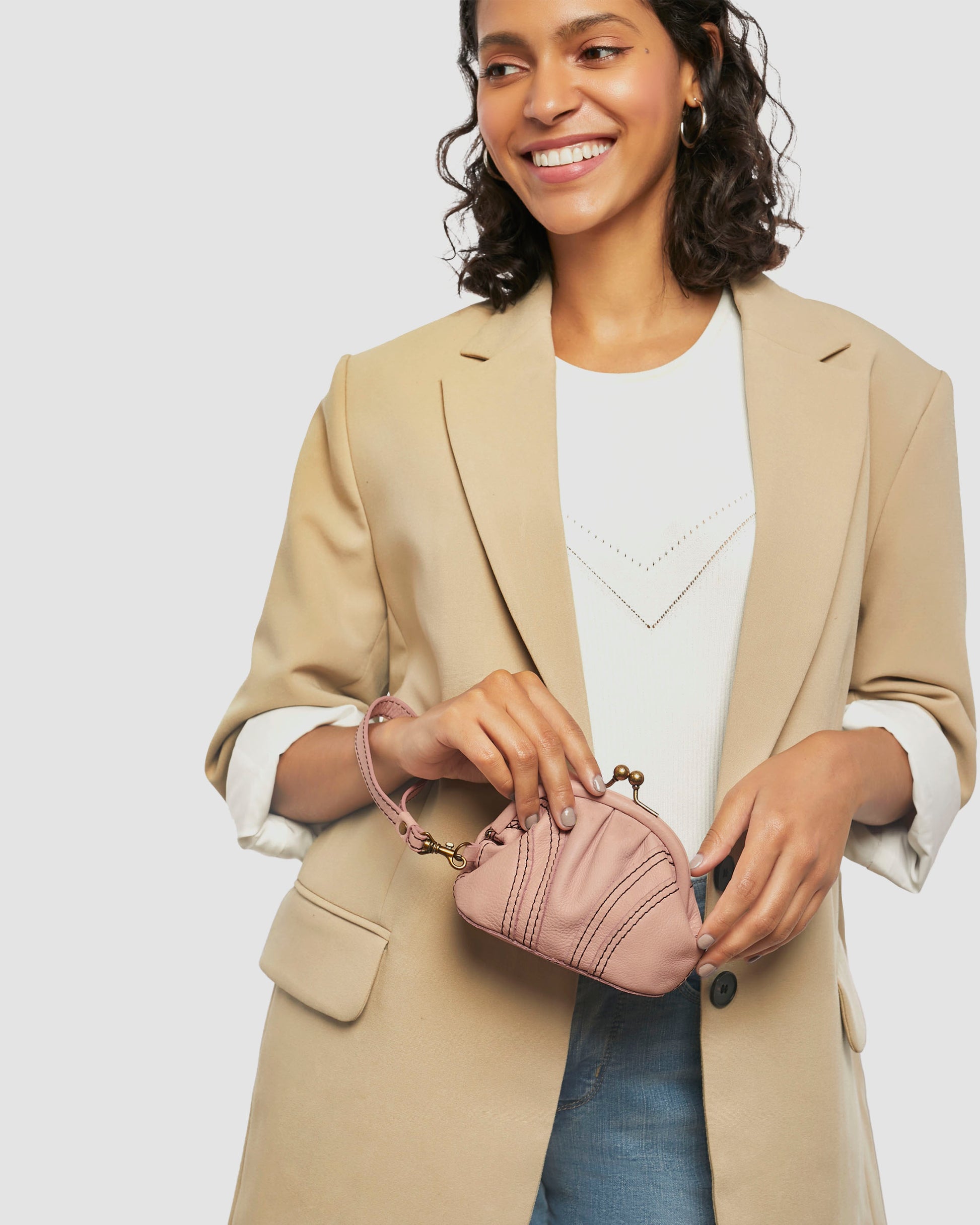 A person beams in a beige blazer over a white top and blue jeans, holding an American Leather Co. Dora Frame Coin Wristlet that perfectly complements their look.