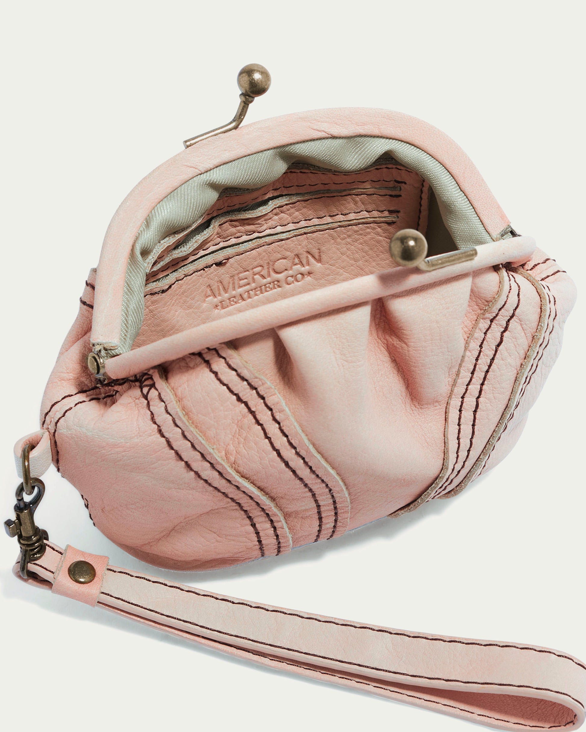 The Dora Frame Coin Wristlet by American Leather Co. is a small light pink leather handbag with metal clasps, revealing a light gray interior embossed with the brand name. Crafted from genuine glove leather, it includes a detachable strap and decorative stitching for timeless elegance.