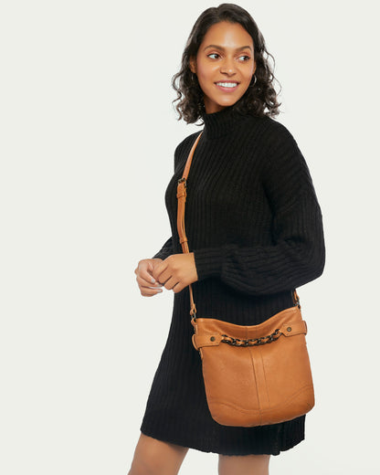 A woman in a black sweater dress smiles while holding the Elton Crossbody by American Leather Co. This vintage bag is crafted from genuine American leather, featuring a decorative chain and antiqued hardware. Her dark, wavy hair accentuates her elegant look against a light background.