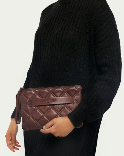 An individual in a black turtleneck sweater is holding the Emerson Quilted Wristlet by American Leather Co., a brown quilted leather clutch with a wrist strap.