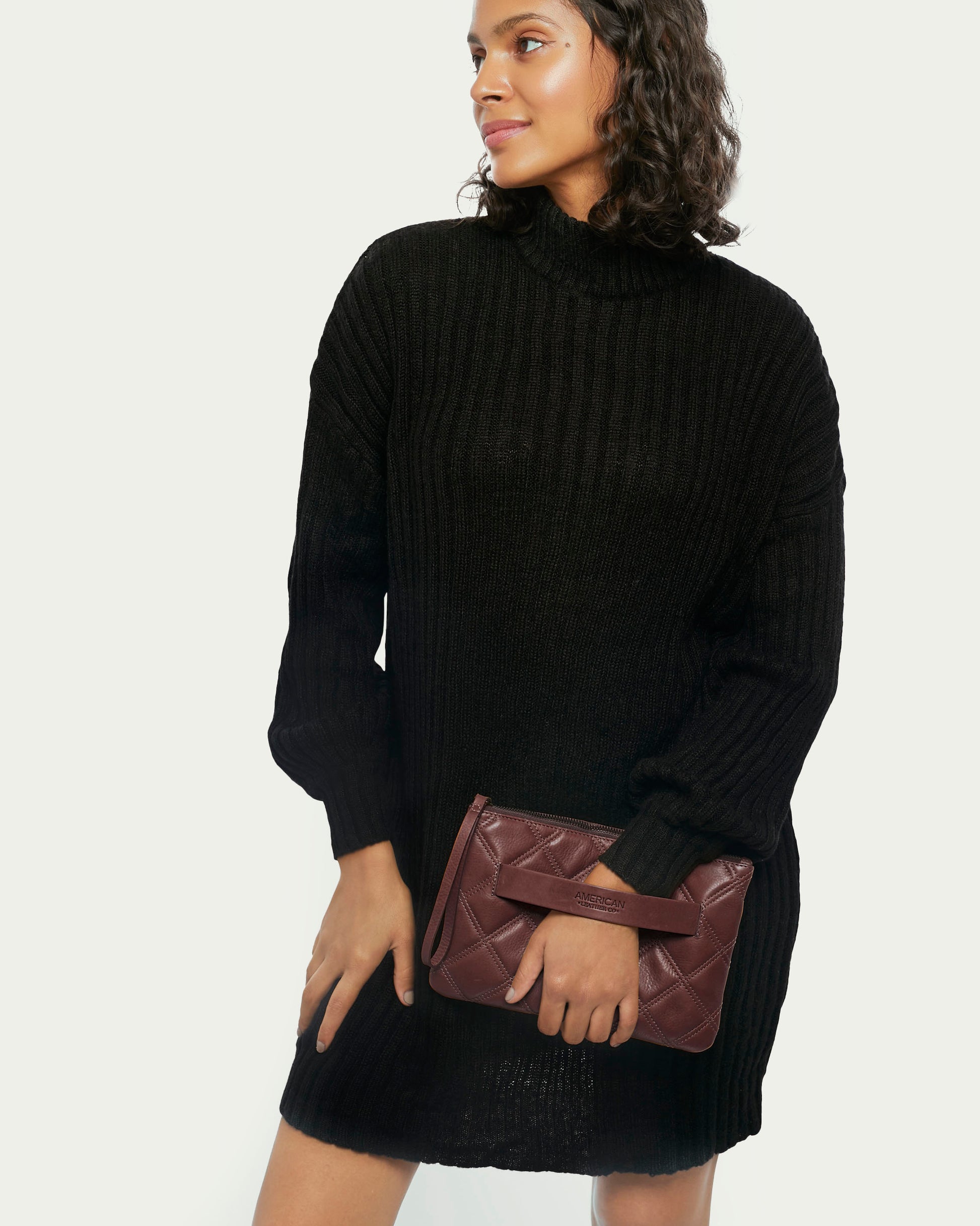 Wearing a black turtleneck sweater dress, a person holds the Emerson Quilted Wristlet from American Leather Co. They have curly hair and are smiling while looking to the side against an off-white background.