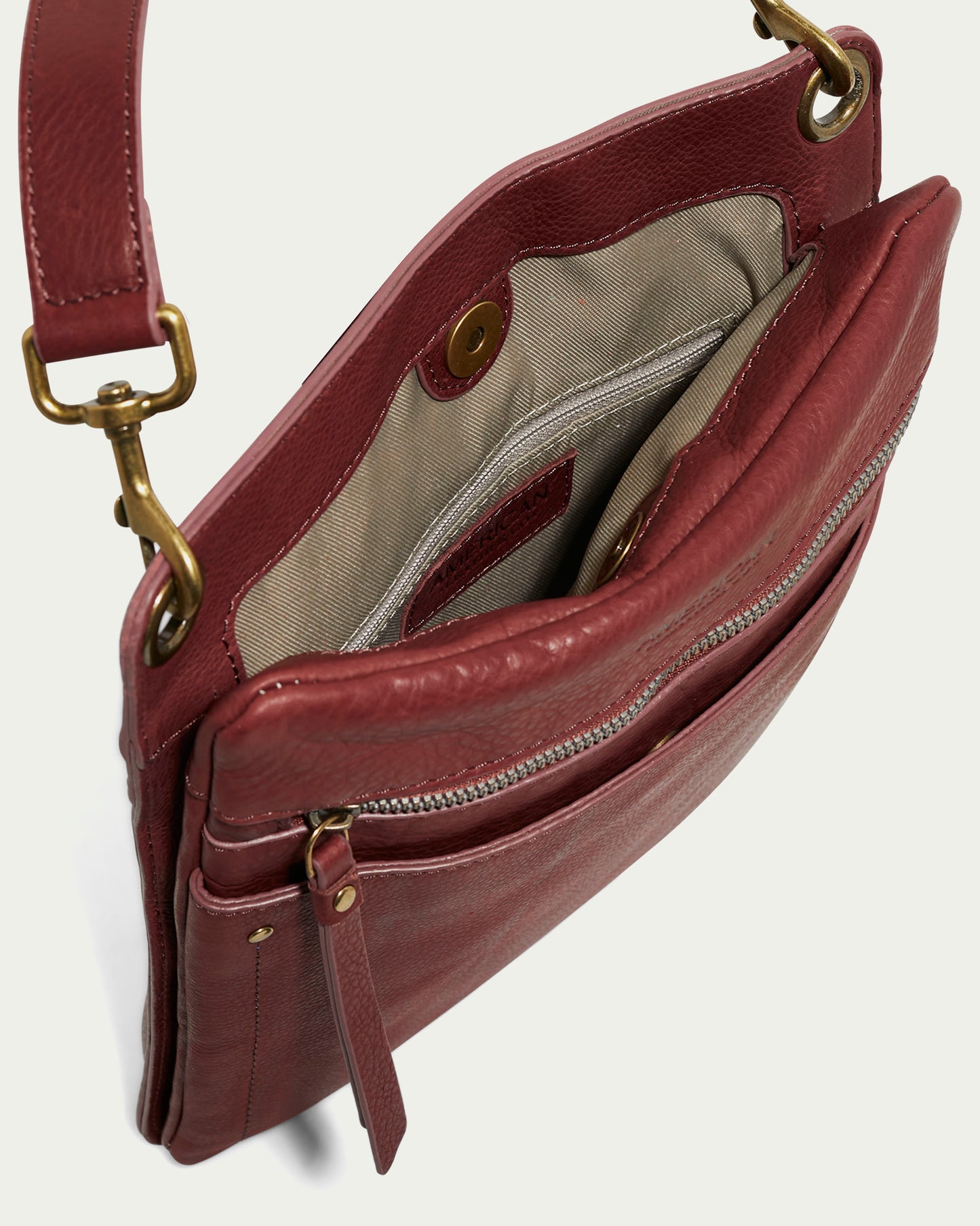 The Lily Multi Compartment Crossbody by American Leather Co. is a red leather handbag with an open top, beige lining, zipper pocket, snap button closure, brass clasp strap, and an external front zipper pocket.