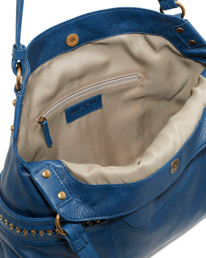 The Sumner Satchel by American Leather Co. is a fashionable accessory crafted from genuine American leather in a striking blue color, complemented by a beige interior lining. This chic bag includes a zipped pocket and decorative exterior studs, combining practicality with style.