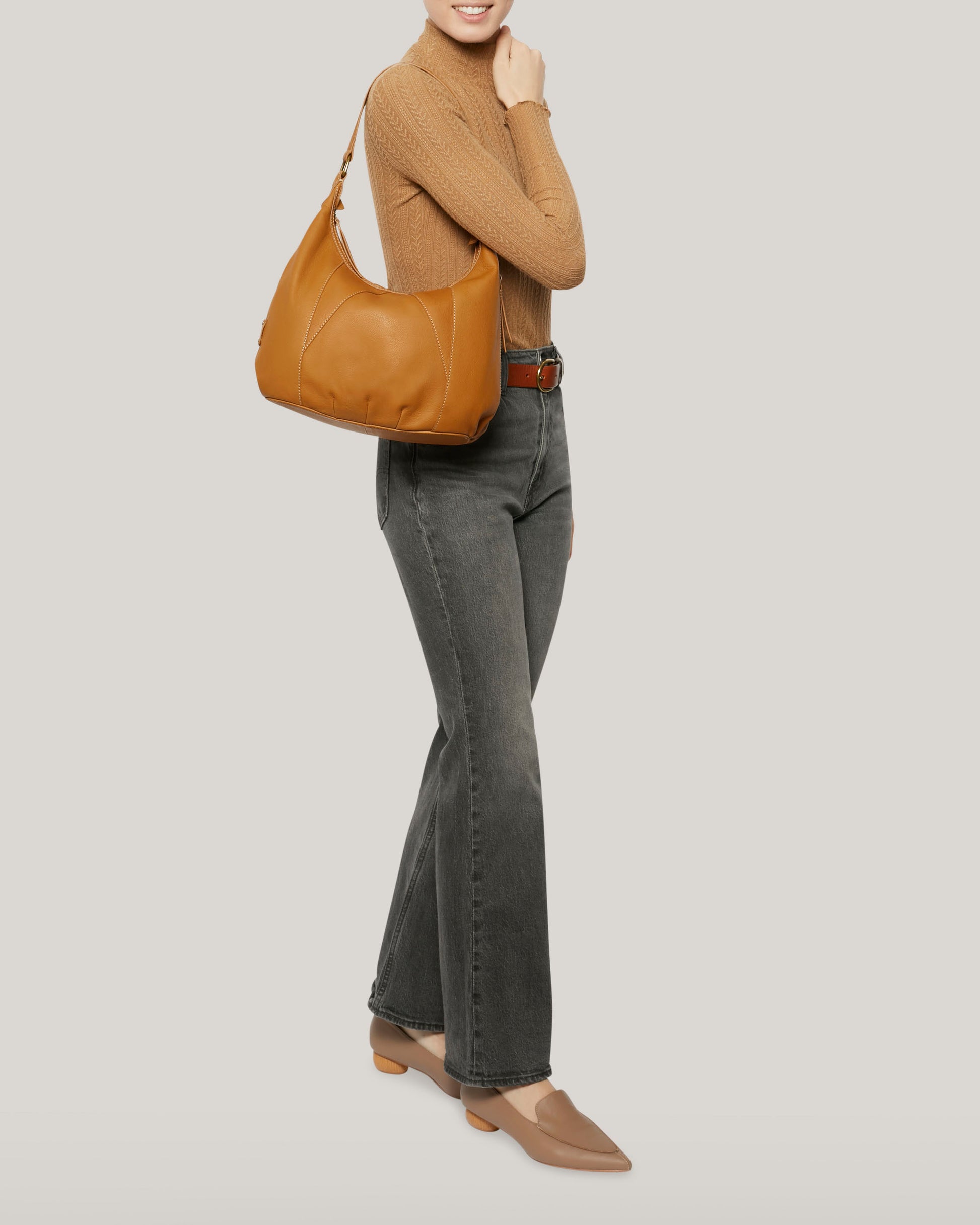 A person with timeless style is dressed in a brown turtleneck sweater, dark grey jeans, and tan loafers. They pose with the Vino Bucket Hobo from American Leather Co., a large leather hobo bag crafted from genuine American leather, against a light grey background.