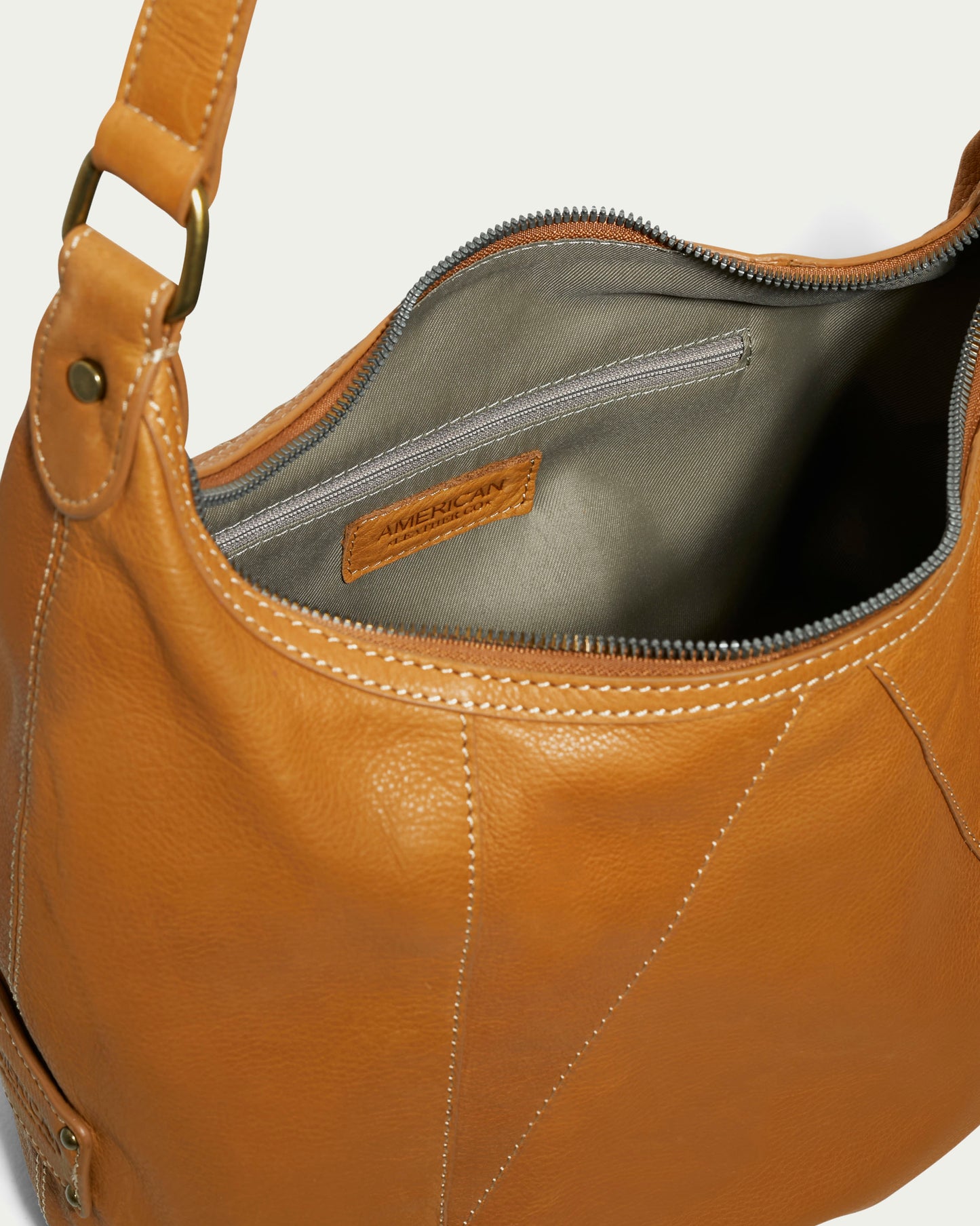 The Vino Bucket Hobo by American Leather Co. is a classic accessory crafted from genuine American leather in a rich brown hue. It features a shoulder strap and opens to reveal a gray fabric interior, complete with a zippered pocket for added convenience. The design is accented with brass hardware and visible stitching, while the interior hosts a thoughtfully sewn label with text.