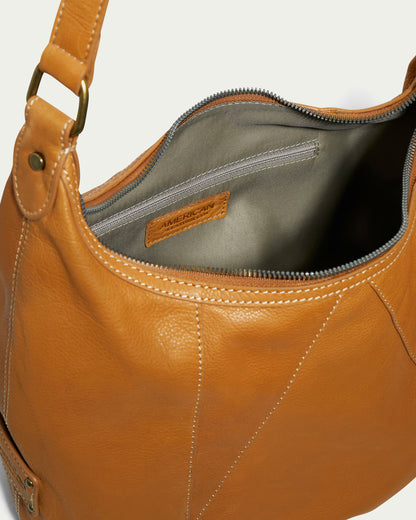 The Vino Bucket Hobo by American Leather Co. is a classic accessory crafted from genuine American leather in a rich brown hue. It features a shoulder strap and opens to reveal a gray fabric interior, complete with a zippered pocket for added convenience. The design is accented with brass hardware and visible stitching, while the interior hosts a thoughtfully sewn label with text.