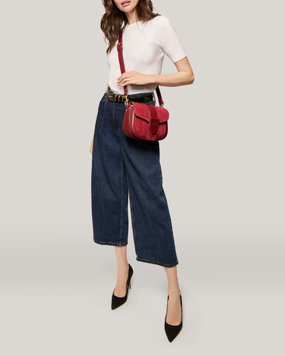 A person with wavy hair wears a white short-sleeve top, high-waisted wide-leg jeans, and black pointed-toe heels. They carry an American Leather Co. Aiken Flap Crossbody, a small red genuine leather bag with a front flap buckle. The background is a plain, light grey color.