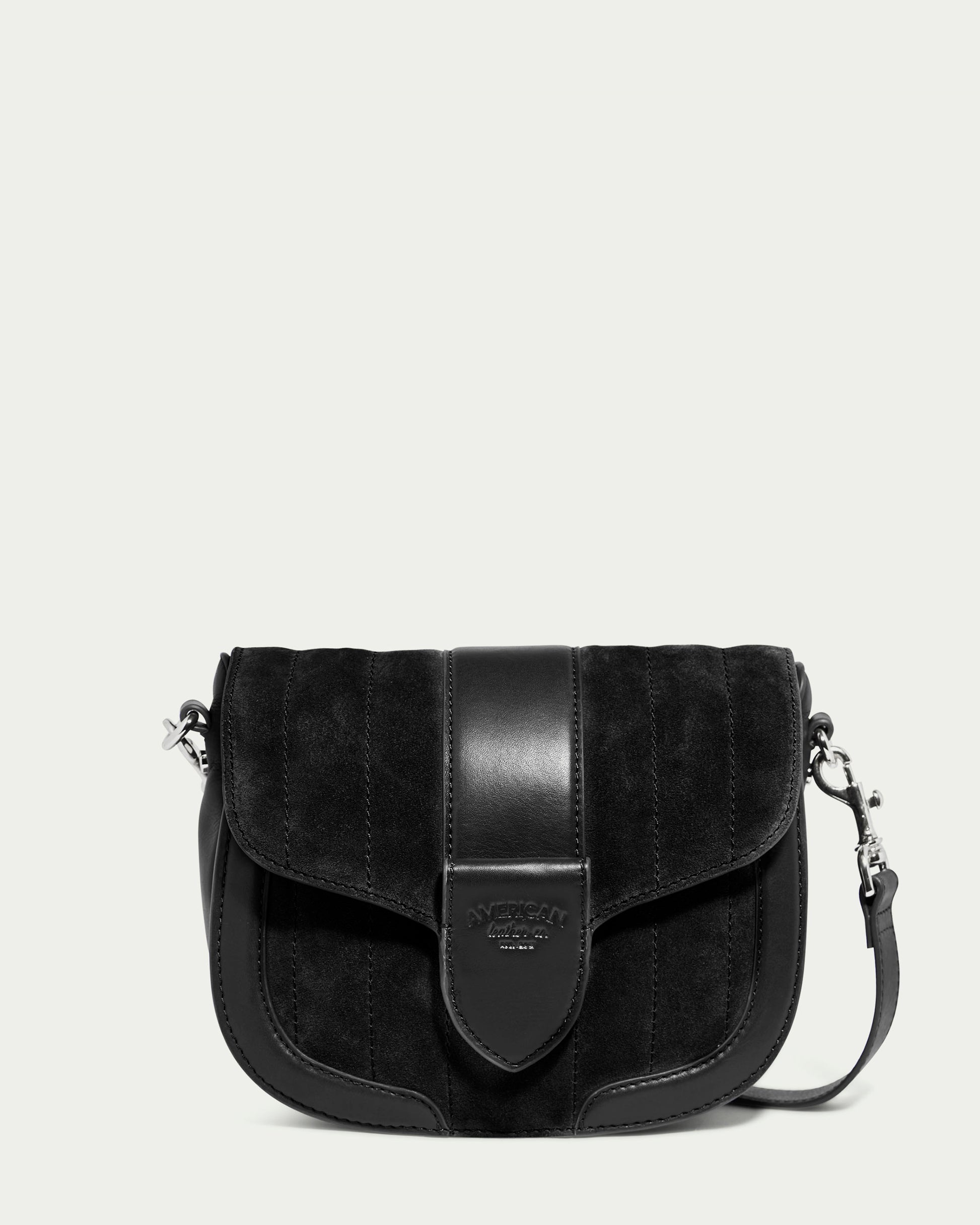 The Aiken Flap Crossbody from American Leather Co. is a black suede and genuine leather bag with a front flap closure. The flap is adorned with a vertical leather strip featuring an embossed logo patch at the center. Accentuated by silver-tone hardware, this sophisticated bag includes an adjustable, detachable shoulder strap with clasps on both sides.