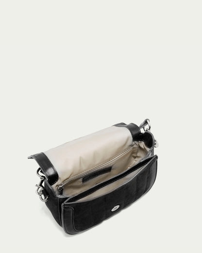 The Aiken Flap Crossbody by American Leather Co. is shown open, revealing its cream-colored interior. The black genuine leather purse features a silver clasp on the front, silver hardware on the sides, and an interior zippered pocket along with an additional open pocket. It boasts suede detailing and comes with a detachable strap. The background is plain white.