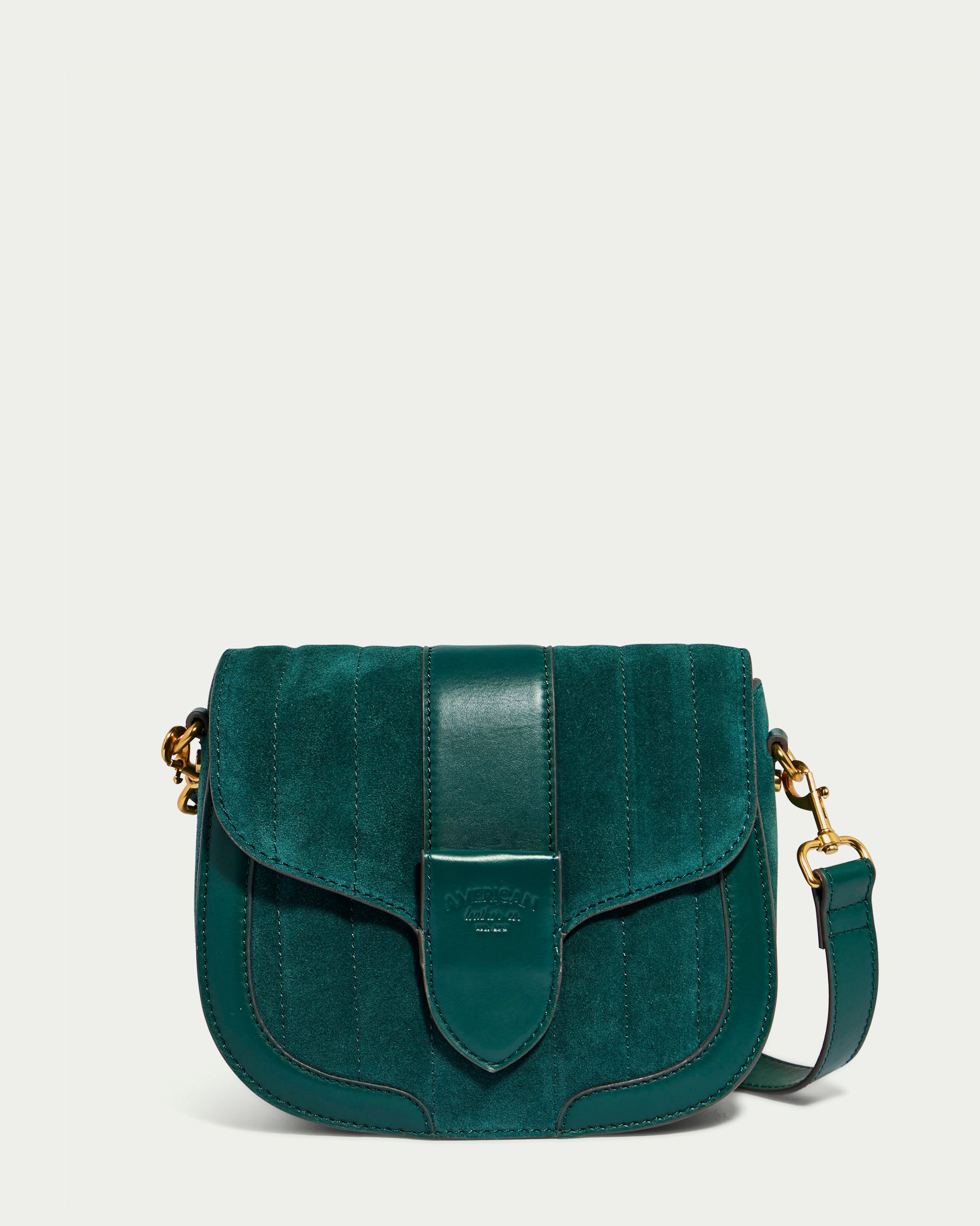 The Aiken Flap Crossbody by American Leather Co. is a genuine leather bag in dark green with a suede and leather finish, featuring a front flap closure, decorative vertical stitching, and gold-tone hardware. The bag has an adjustable strap and the brand's logo embossed on the front flap.