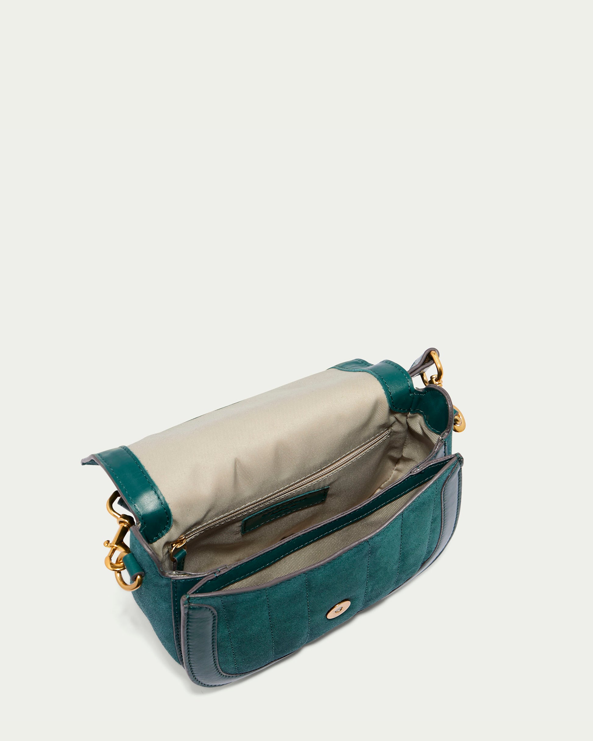 An image of the Aiken Flap Crossbody from American Leather Co. is shown. This green rectangular shoulder bag features a flap opening and is crafted from genuine leather with both leather and suede textures on the exterior. Inside, it has a beige fabric-lined interior complete with a zippered pocket, and it closes securely with a gold clasp. The adjustable strap attachments are accented with gold hardware.