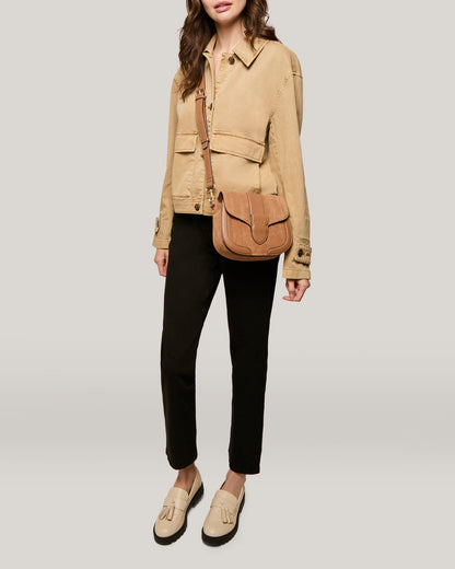 A person is shown wearing a beige jacket, black pants, and beige loafers. They are carrying the American Leather Co.'s Aiken Flap Crossbody, a tan leather bag with suede detailing. The background is plain and light gray. The person's head is cropped out of the image.