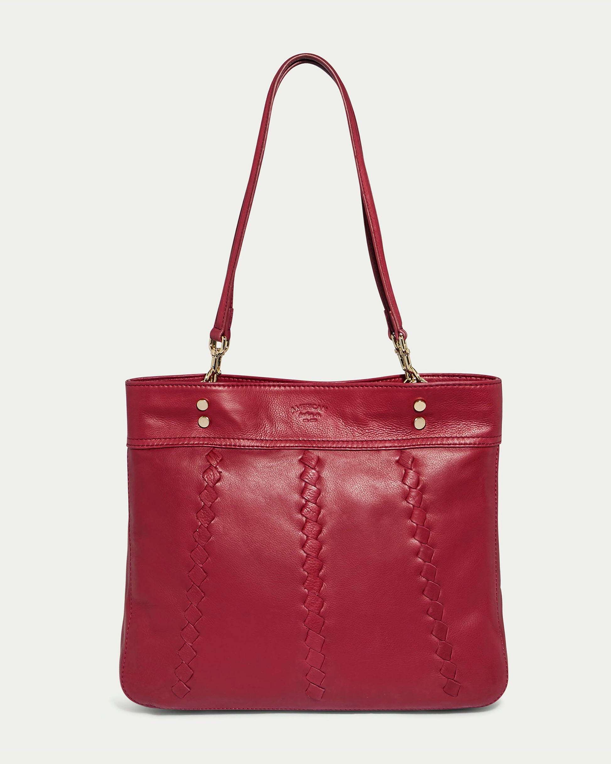 The Alpine Triple Compartment Tote by American Leather Co. is a stylish red handbag crafted from genuine leather, featuring braided detailing on the front and gold-tone hardware at the strap attachments. The bag showcases two parallel shoulder straps, a flat bottom for stability, and a triple compartment design for optimal organization. Perfect for everyday use or special occasions.