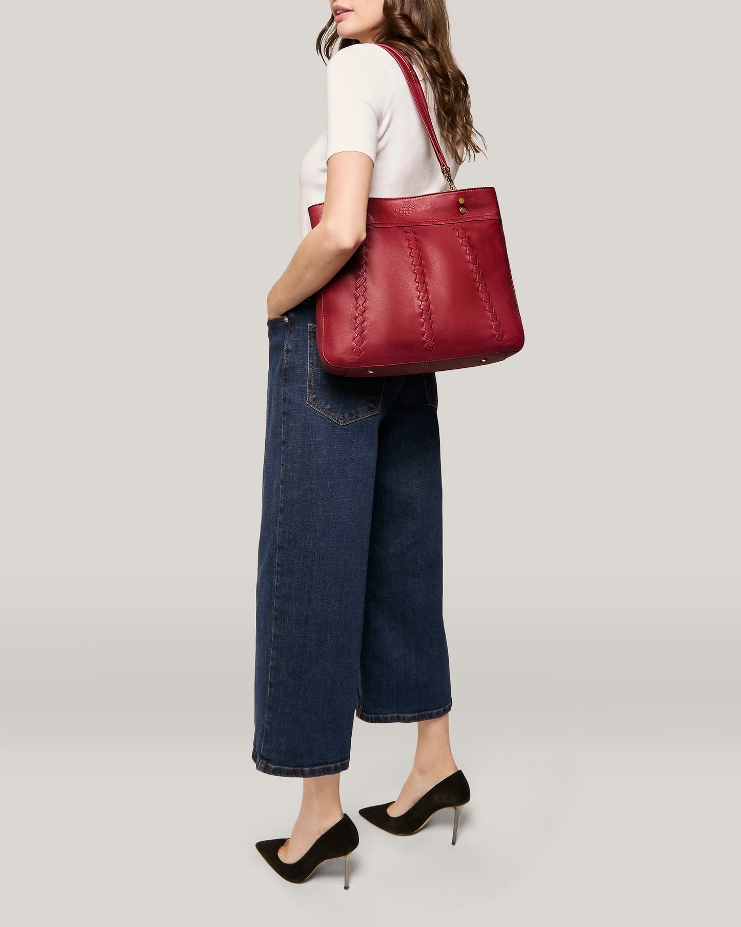 A woman stands in a relaxed pose wearing a white short-sleeve top, high-waisted wide-leg blue jeans, and black high-heeled shoes. She has the Alpine Triple Compartment Tote by American Leather Co., a red genuine leather handbag with three organizational compartments, slung over her shoulder. The background is a plain light grey.