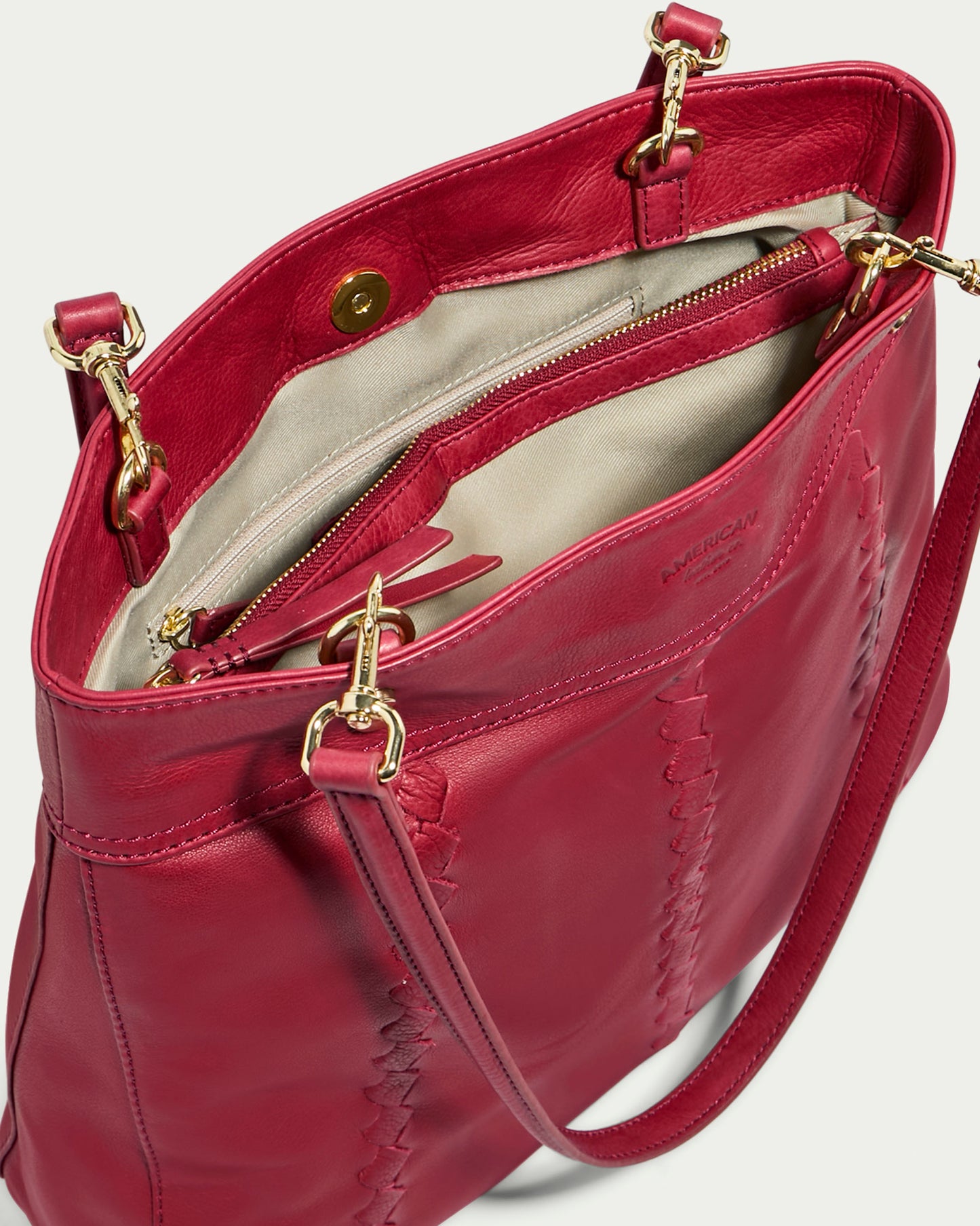 The Alpine Triple Compartment Tote by American Leather Co. is a stylish red handbag crafted from genuine leather, featuring gold hardware. The open bag reveals its beige interior lining and includes a zippered inner pocket within its triple compartment design. Decorative vertical stitching adorns the exterior of the bag, which also comes with a detachable shoulder strap for versatile use.