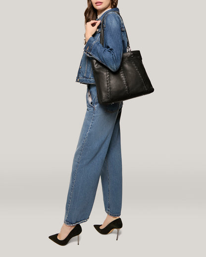 A person wearing a denim jacket and jeans is shown from the side, holding an American Leather Co. Alpine Triple Compartment Tote with a genuine leather exterior over their shoulder. They are also wearing black high-heeled shoes. The background is a plain, light grey color.