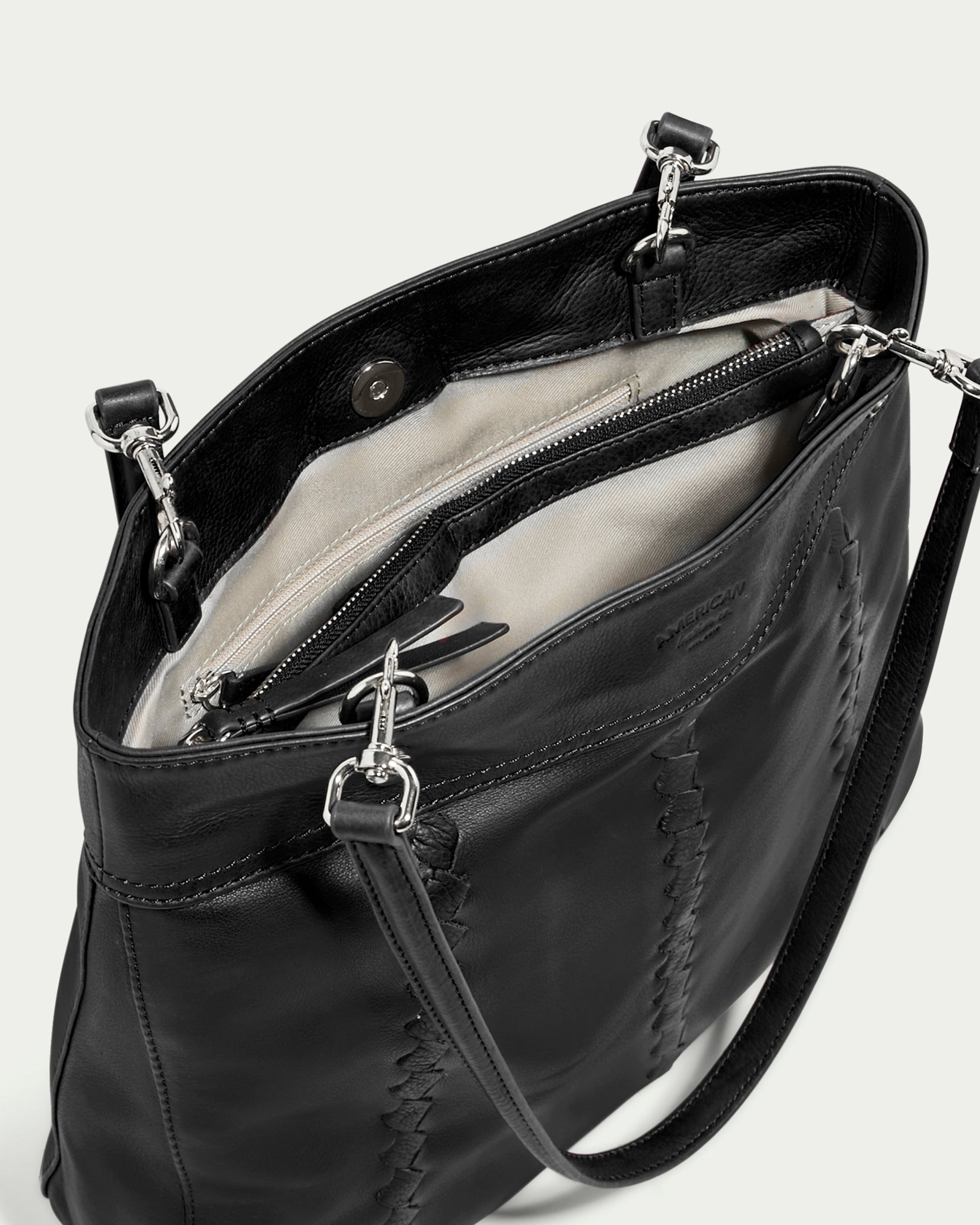 Introducing the American Leather Co. Alpine Triple Compartment Tote: a sleek black leather tote bag featuring elegant silver hardware and a long strap. Crafted from genuine leather, its exterior opens to reveal a spacious interior lined with beige fabric, complete with a zippered compartment and an inner pocket for meticulous organization. The bag showcases an open design that highlights its thoughtfully designed triple compartments.