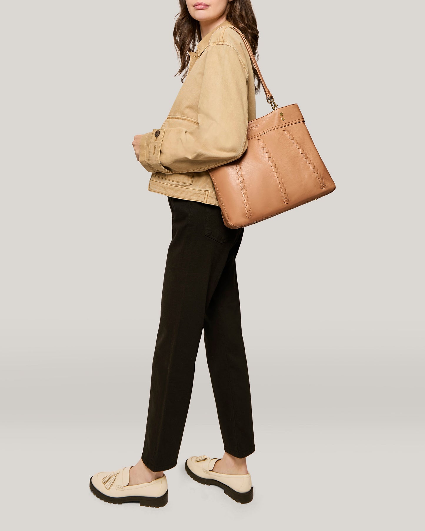 A person wearing a beige jacket, black pants, and cream-colored loafers with bows on top is carrying an Alpine Triple Compartment Tote by American Leather Co., slung over one shoulder. They are standing against a plain, light gray background. Only their midsection and legs are visible, with their face out of frame.