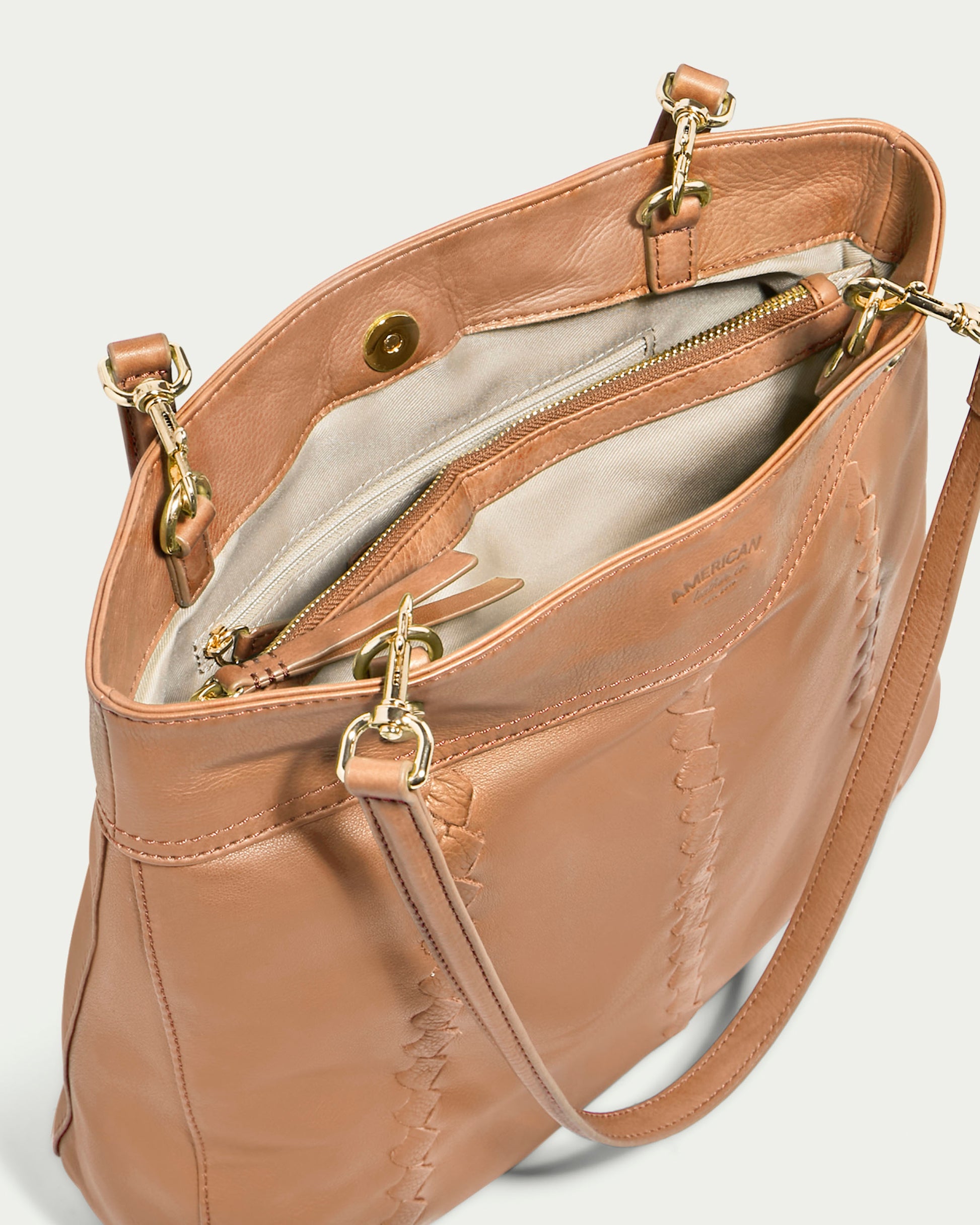 The Alpine Triple Compartment Tote by American Leather Co. is a tan genuine leather handbag featuring gold hardware and an open top. Inside, it boasts three compartments, including one with a zipper. The exterior showcases intricate stitching details, and the bag comes with a detachable shoulder strap equipped with gold clasps.