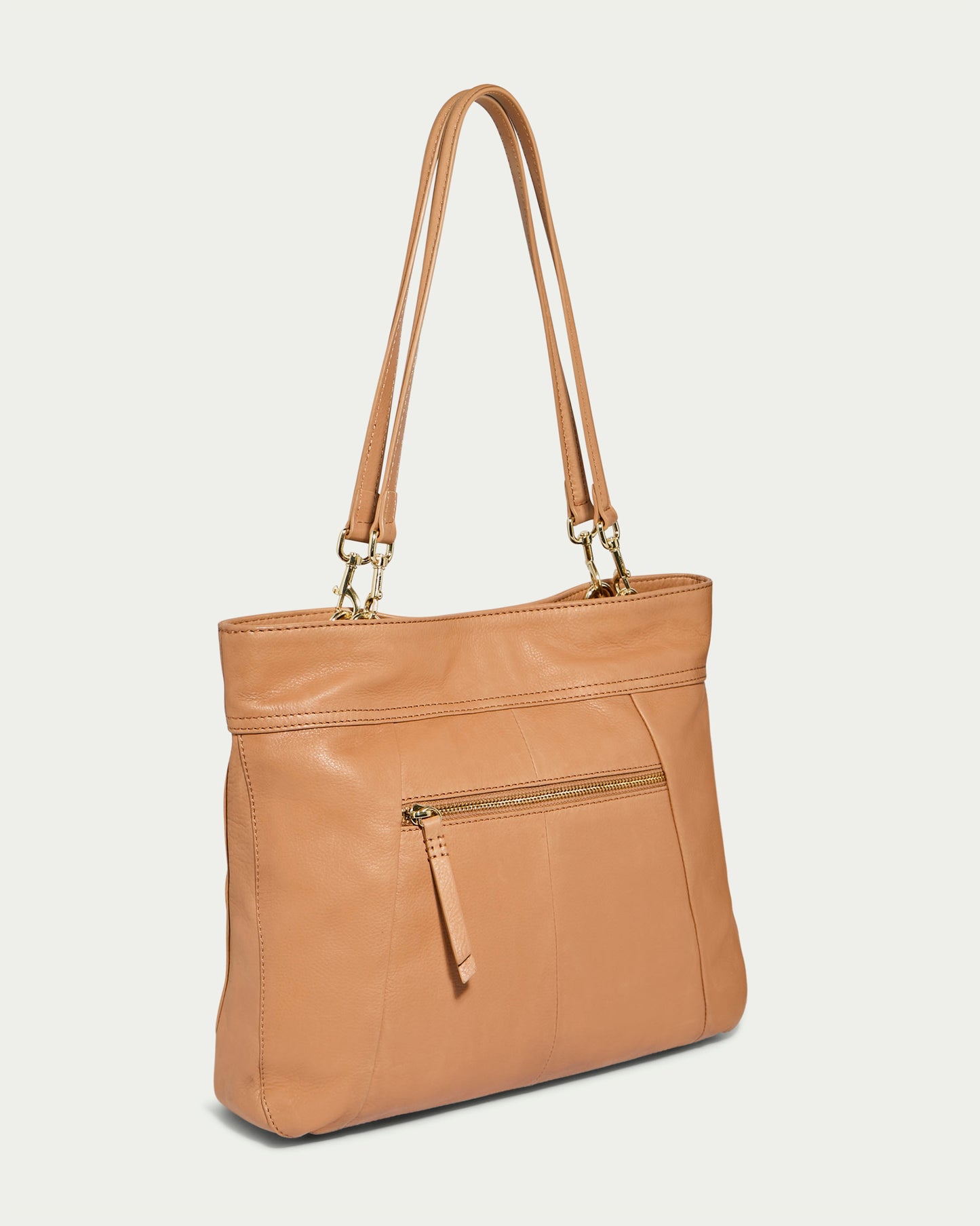 The Alpine Triple Compartment Tote from American Leather Co. is a tan, genuine leather handbag with two shoulder straps and gold hardware. It features a front zippered pocket with a leather pull tab and boasts a simple, sleek design, making it perfect as a laptop bag. The background is off-white.