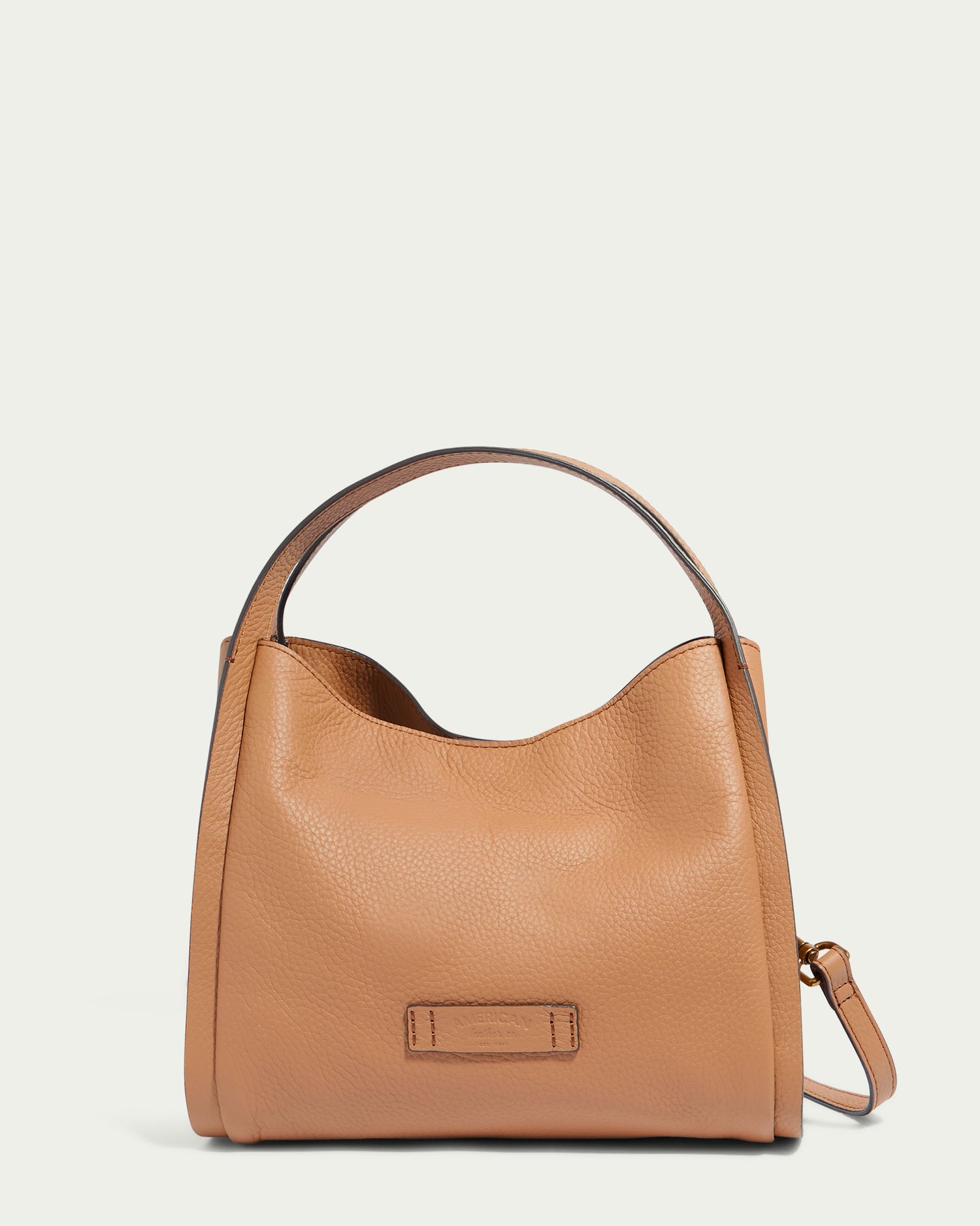 The Auburn Triple Compartment Convertible Crossbody by American Leather Co. is a tan handbag made from genuine leather with a pebble grain texture. It boasts a short, wide handle, an open-top with a slightly curved design, and a small rectangular leather patch on the front. Additionally, it comes with a detachable crossbody shoulder strap attached to the sides.