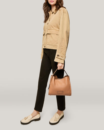 A woman stands against a plain background, wearing a beige jacket and black pants. She holds the Auburn Triple Compartment Convertible Crossbody by American Leather Co., which is made of genuine leather and weatherproof with a short handle. She wears beige loafers, her brown hair cascades over her shoulders, and her face is partially obscured.