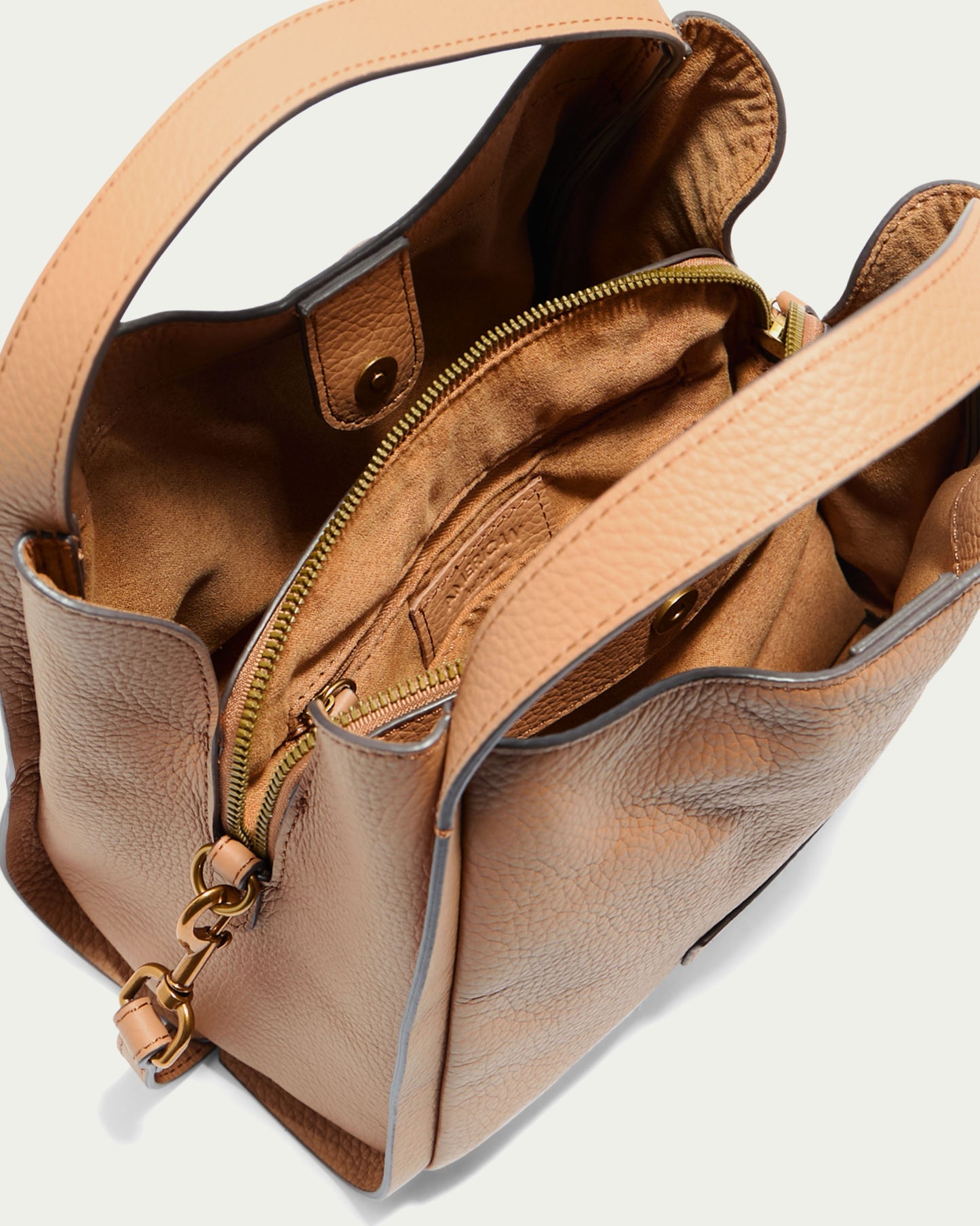 A top-down view of the American Leather Co. Auburn Triple Compartment Convertible Crossbody shows its open tan pebbled leather design. Crafted from genuine leather, the interior showcases a central zippered compartment, brown lining, and an additional open pocket. Snap closures secure the top edges, while gold-tone hardware accents this versatile crossbody style.