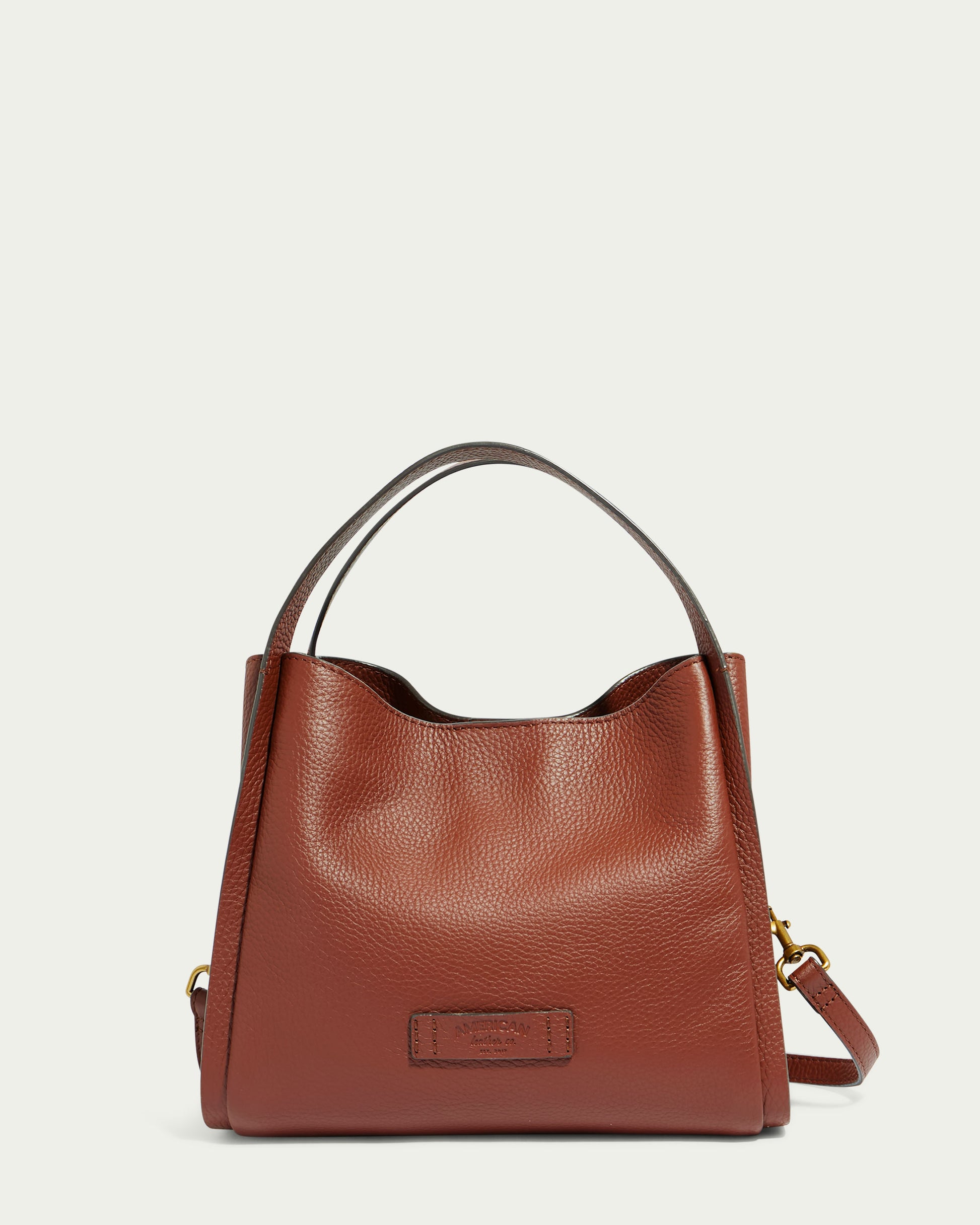 Introducing the Auburn Triple Compartment Convertible Crossbody by American Leather Co. — a brown genuine leather handbag with a textured surface, featuring two short handles. It showcases a small rectangular patch on the front with subtle branding. The bag also includes a detachable shoulder strap adorned with gold-tone hardware accents. The background is plain white.