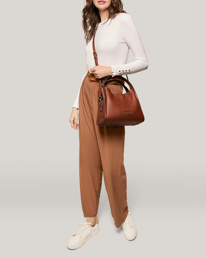 A person with long hair is wearing a white long-sleeved top, brown high-waisted pants, and white sneakers. They are carrying the Auburn Triple Compartment Convertible Crossbody bag from American Leather Co., which features a genuine leather design and a top handle. The background is plain light gray.