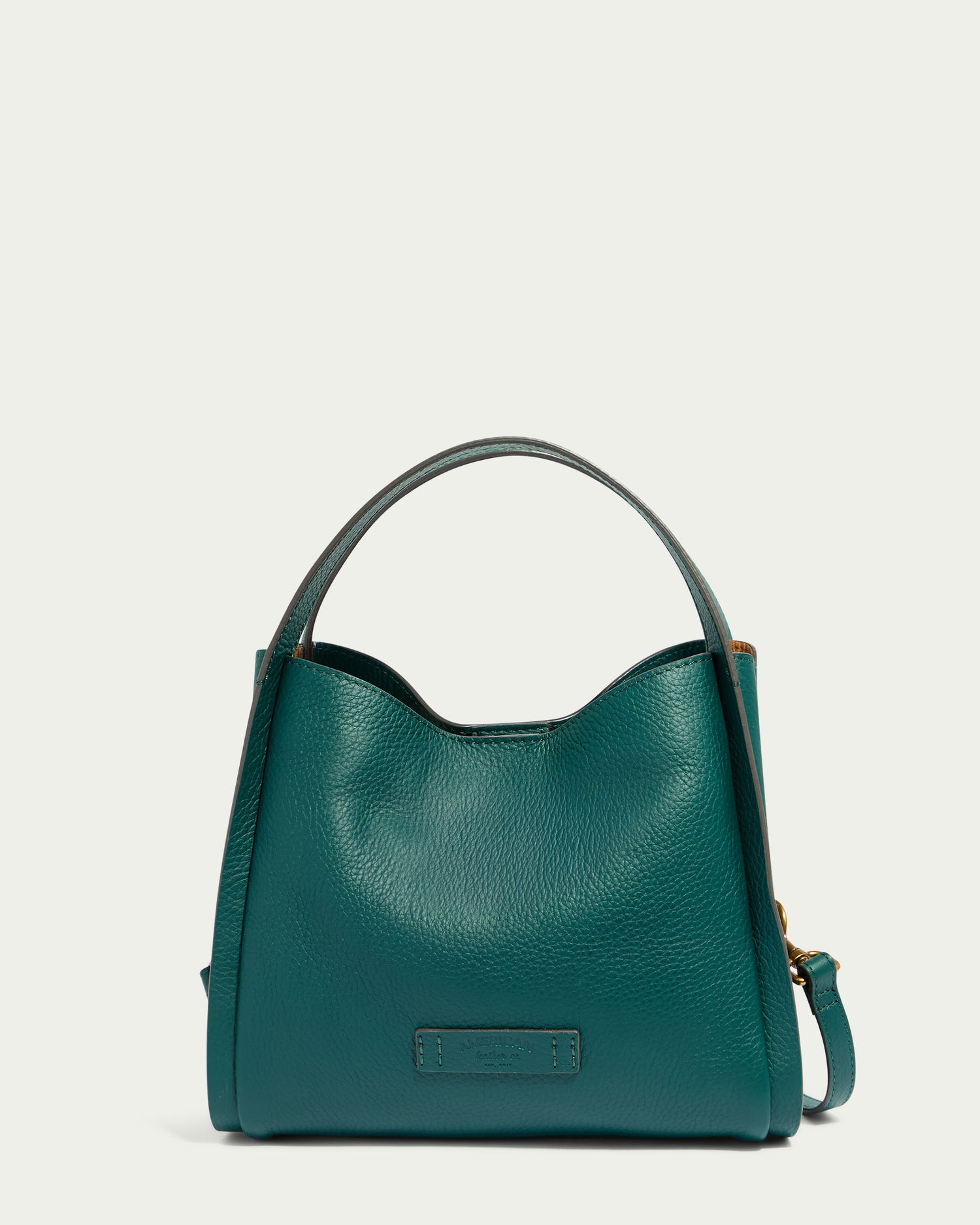 Introducing the Auburn Triple Compartment Convertible Crossbody by American Leather Co. This genuine leather handbag comes in a striking deep teal shade, featuring both a shoulder strap and a short top handle for versatile wear. The bag has an elegant, slightly slouchy shape complemented by minimal gold hardware. It boasts triple entry compartments and is adorned with a small rectangular logo patch on the front. The background is set against a light, neutral color to highlight its beauty.