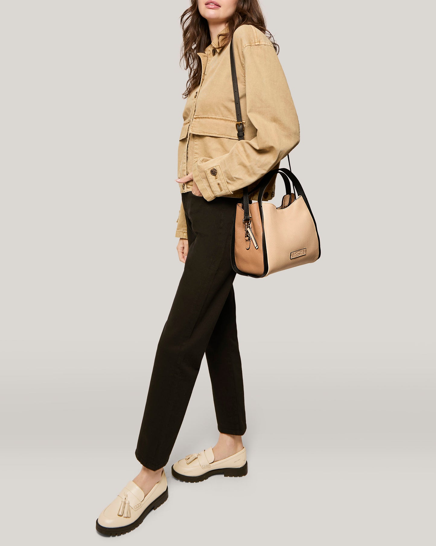 A stylish person wears a beige jacket, black pants, and beige loafers. They are holding an American Leather Co. Auburn Triple Compartment Convertible Crossbody, a genuine leather handbag in beige and black, by its handles with the black crossbody shoulder strap attached. The background is neutral gray.